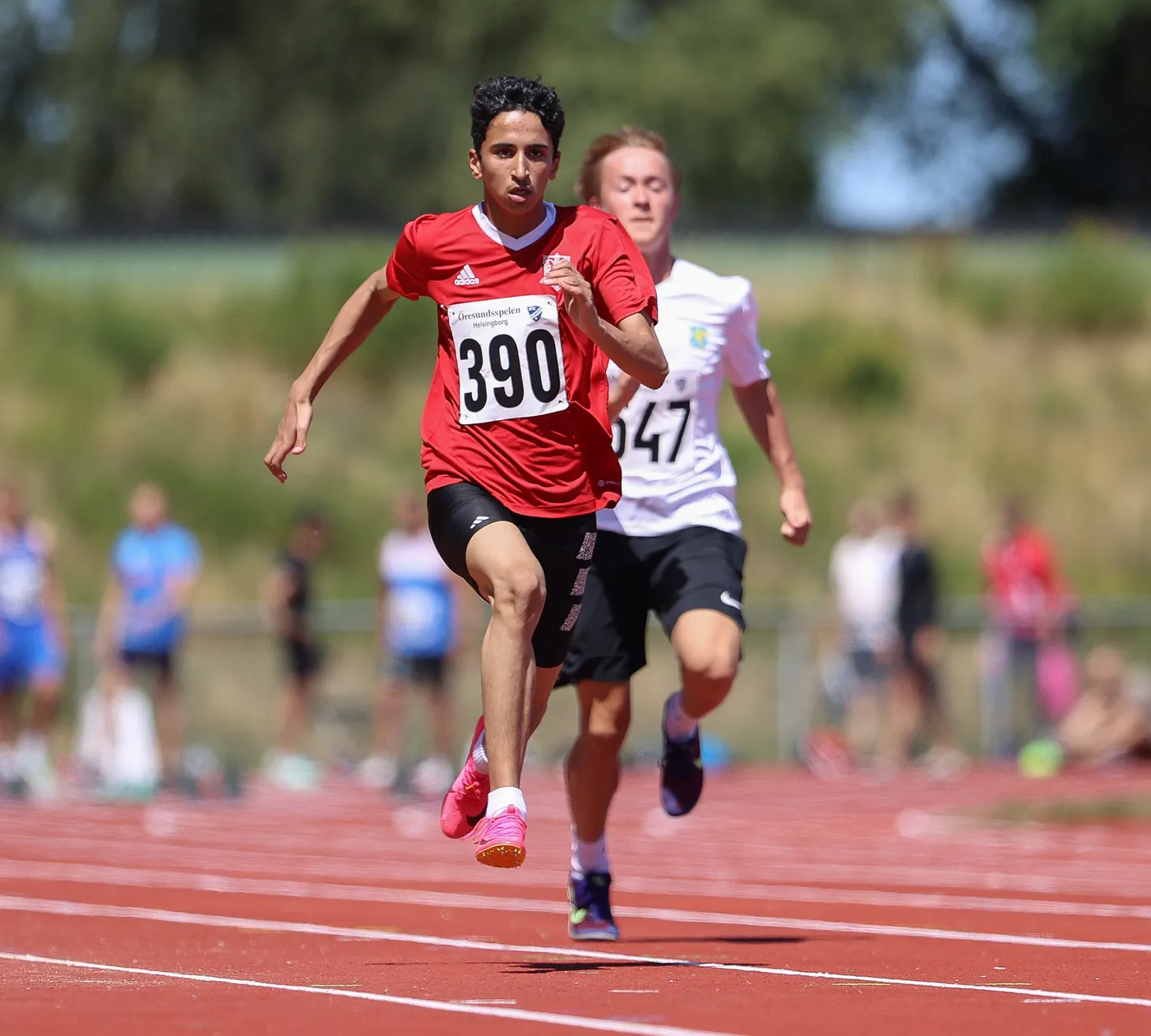 شاب يجري في سباق على المضمار، يرتدي قميصًا أحمر يحمل الرقم 390، بينما يتقدم على متسابق آخر يرتدي قميصًا أبيض يحمل الرقم 647. الخلفية تتضمن حشدًا من المشاهدين واللاعبين في يوم مشمس.