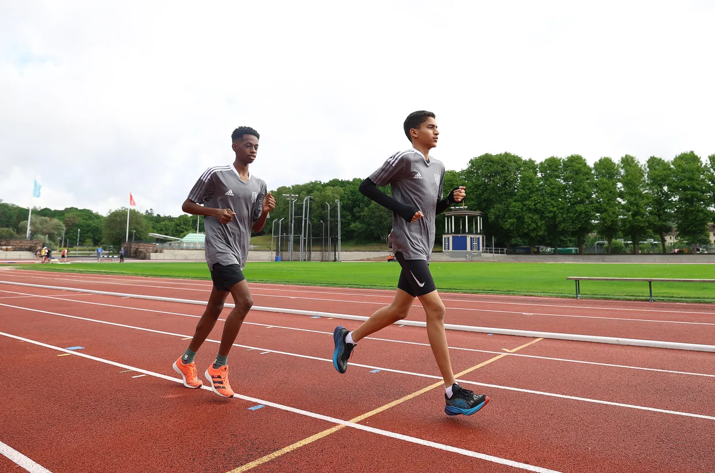 شابين يرتديان قمصان رياضية رمادية وشورتات قصيرة، يجريان على حلبة رياضية. يظهر في خلفية الصورة ملعب أخضر وأعمدة رياضية، بينما السماء مغطاة بسحاب خفيف.