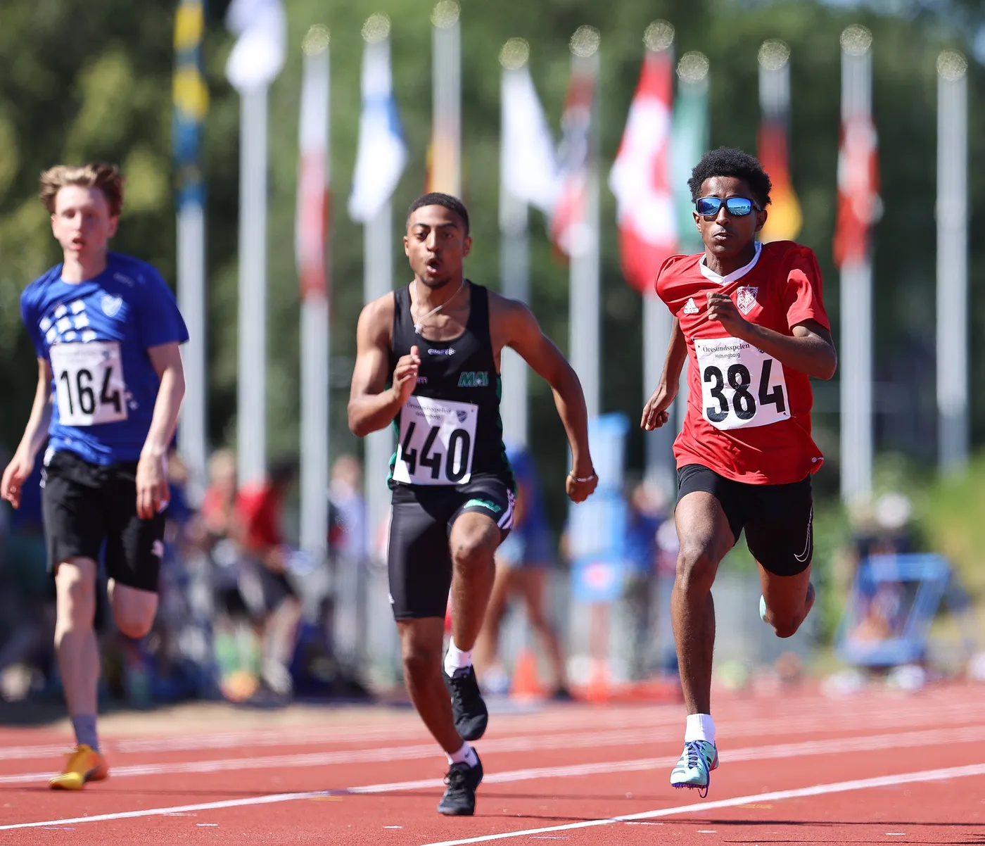 عداءان يتنافسان على مضمار السباق، حيث يرتدي أحدهم قميصاً أزرق والآخر قميصاً أسود مع رقم 440، بينما العداء الثالث في المقدمة يرتدي قميصاً أحمر يحمل الرقم 384. تظهر أعلام عديدة في الخلفية تحت أشعة الشمس الساطعة.