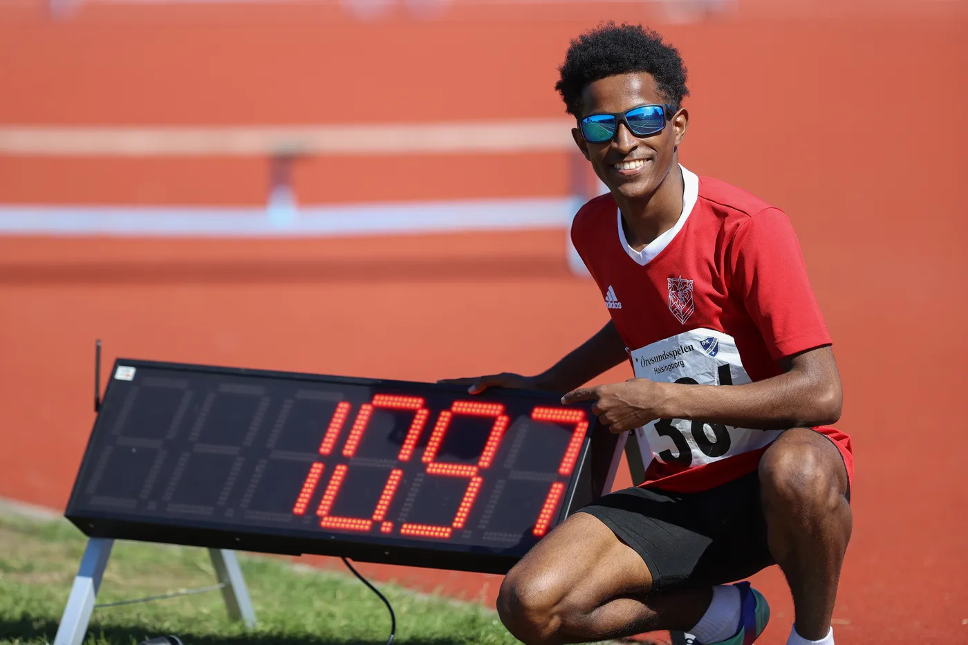 عداء يتبسم ويحمل علامة رقمية توضح توقيت 10.97 ثانية، وهو يرتدي قميصًا أحمر ونظارات شمسية.背景 ملون بالأرضية الحمراء ملعب تتبع حوادث الرياضية.