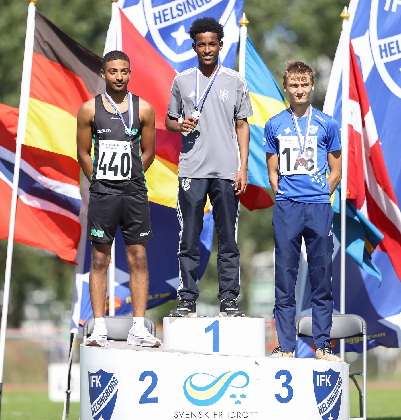 ثلاثة رياضيين يقفون على منصة التتويج، حيث يحمل أحدهم ميدالية فضية ومظهره يشير إلى فوز في سباق. خلفهم أعلام متعددة لألوان البلدان تظهر في الخلفية، بينما يرتدي الرياضيون ملابس رياضية مختلفة، مع شعار يظهر على المنصة.