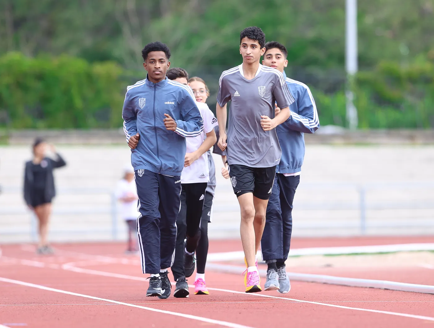 أربعة شبان يجري في حلبة رياضية، يرتدون ملابس رياضية بألوان مختلفة. يظهرون في حالة نشاط وتركيز بينما يتدربون.