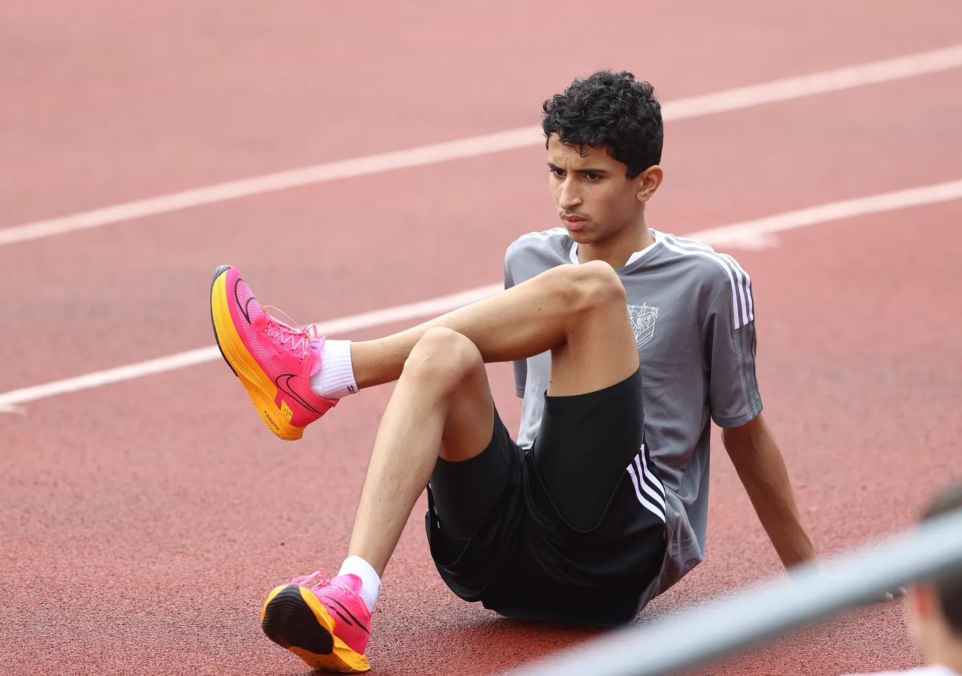 شاب يجلس على مضمار رياضي، يرتدي قميصًا رماديًا وشورتًا أسود مع أحذية رياضية وردية وصفراء. يبدو أنه يفكر أو يستريح بعد ممارسة الرياضة.