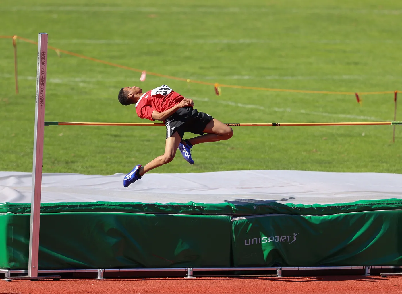 رياضي يتنافس في مسابقة القفز العالي، يقفز فوق العارضة بينما ينحني إلى الوراء. الملعب أخضر، وهناك حصيرة تحت الرياضي لاستقباله بعد القفز.