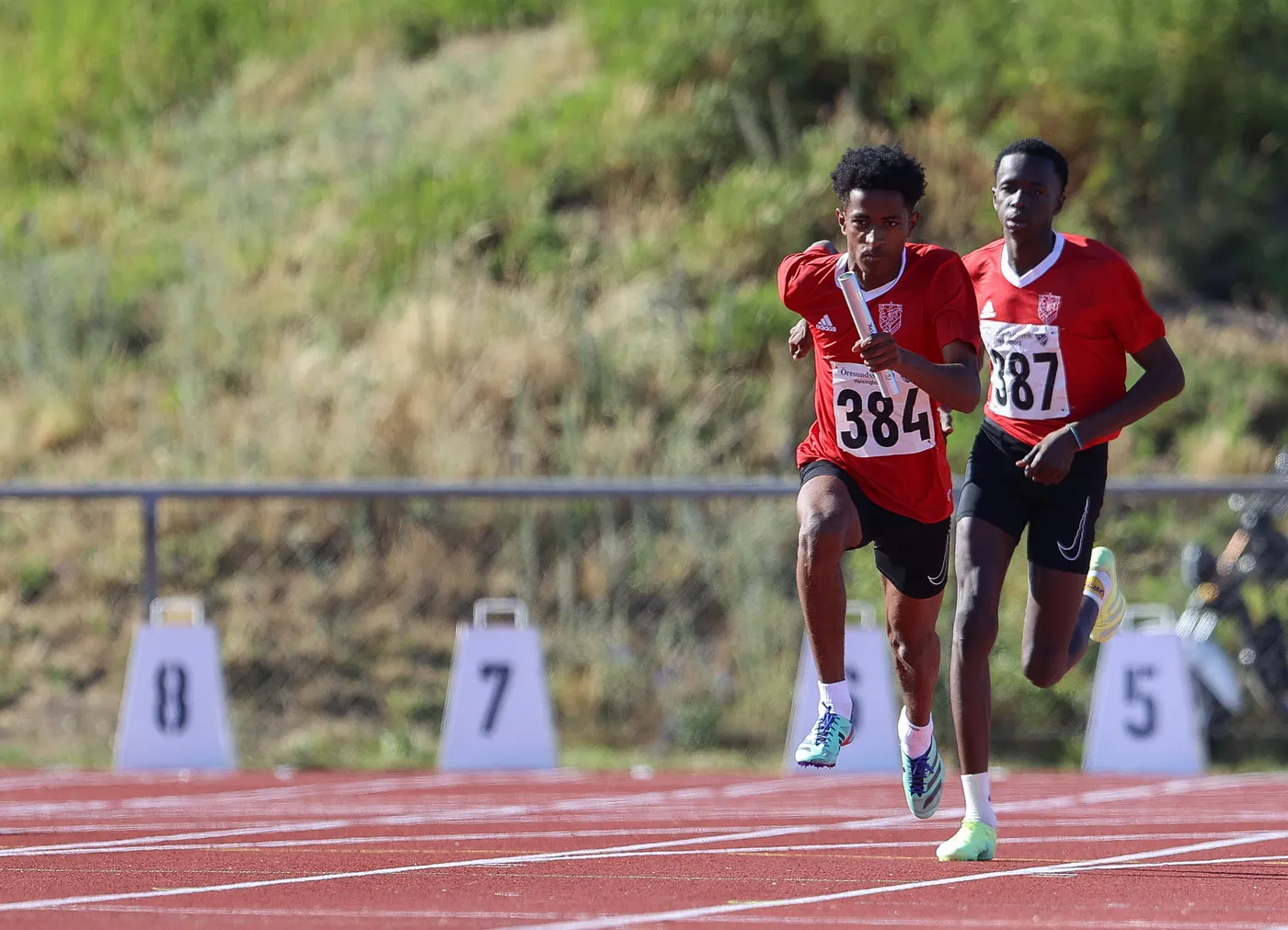 عداءان يرتديان زياً رياضياً أحمر، يركضان على مضمار السباق، بينما يحمل أحدهما عصا التتابع. تظهر الأرقام على صدورهم، مع رقم 384 في المقدمة. خلفهم توجد علامات للزمن على المضمار.