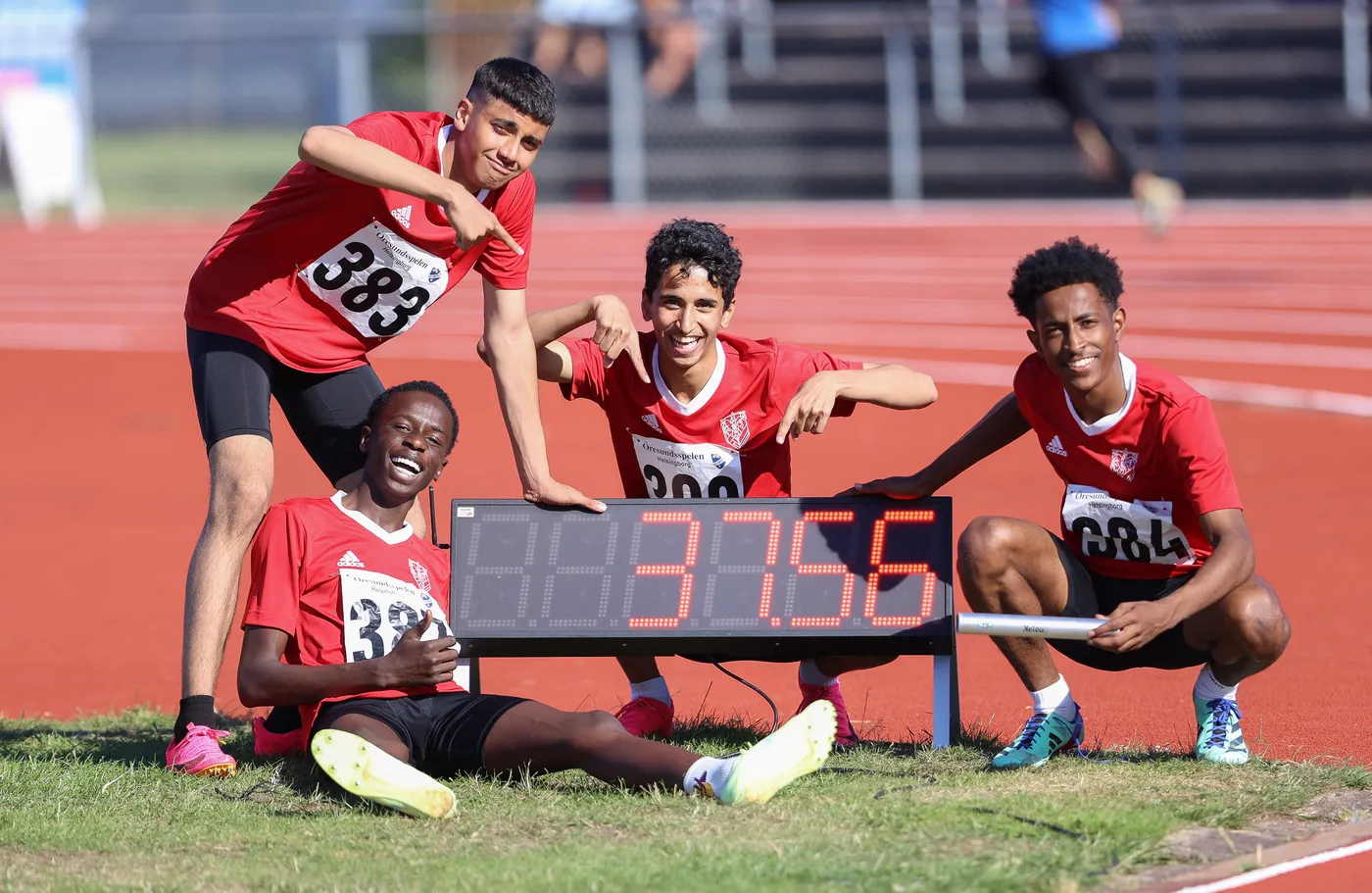 أربعة شباب رياضيين يرتدون قمصان حمراء، يجلسون ويبتسمون بجوار لوحة إلكترونية تظهر وقت 37.56. يقوم أحدهم بالإشارة إلى الرقم بينما يُظهر الآخرون الإشارة بالأصابع، في مضمار رياضي.