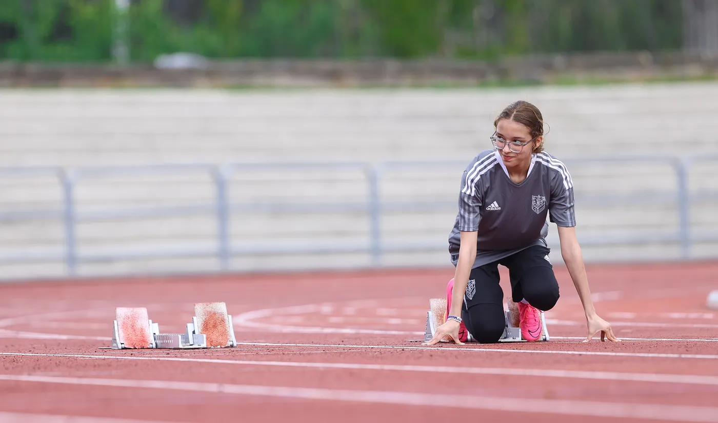 شخص يجلس على مضمار رياضي، يستعد للانطلاق من مخارج البداية. يرتدي ملابس رياضية ويحمل في يده أداة الانطلاق. الجو يبدو هادئاً والخلفية تحتوي على مدرجات فارغة.