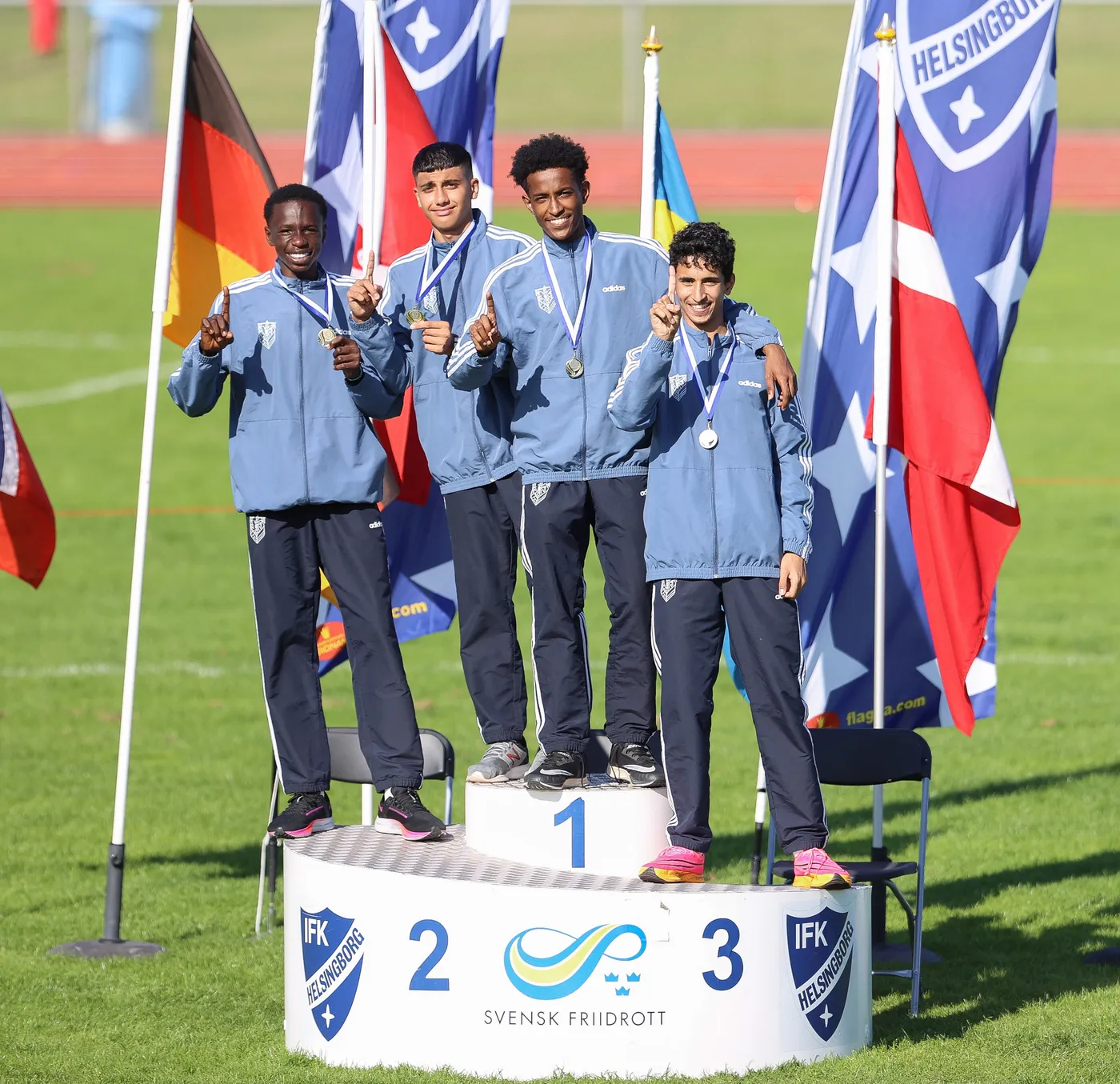 أربعة شبان يقفون على منصة تتويج، يحمل كل منهم ميدالية فضية، مع رفع أحدهم إصبعه إلى الأعلى في إشارة للنجاح. خلفهم ترفرف الأعلام المختلفة، مما يدل على أجواء المنافسة الرياضية.