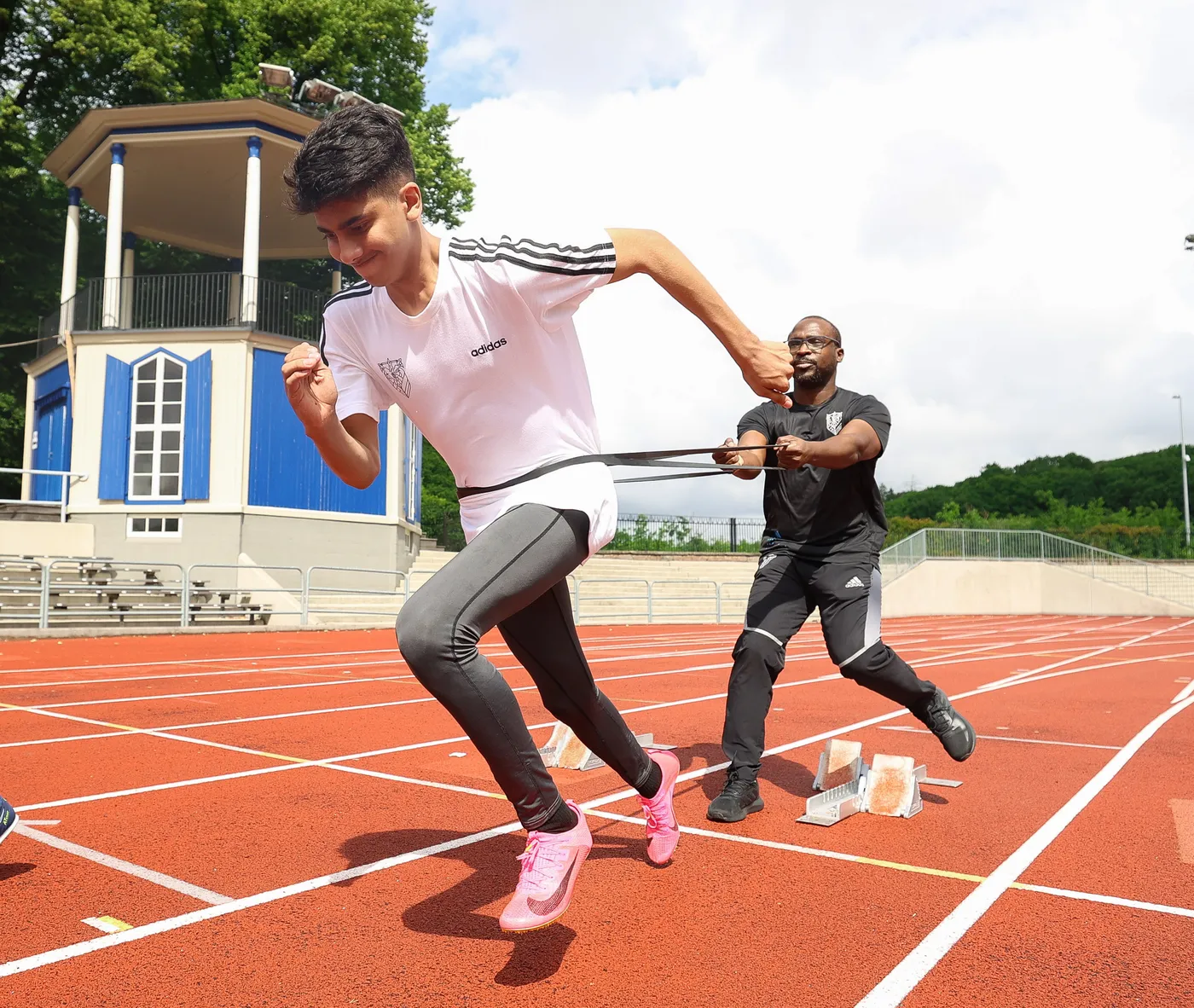 شباب يُمارس رياضة الجري على مضمار رياضي بينما يُساعده مدرب باستخدام حزام لتدريبه. الخلفية تضم مدرجات وزاوية من المبنى.