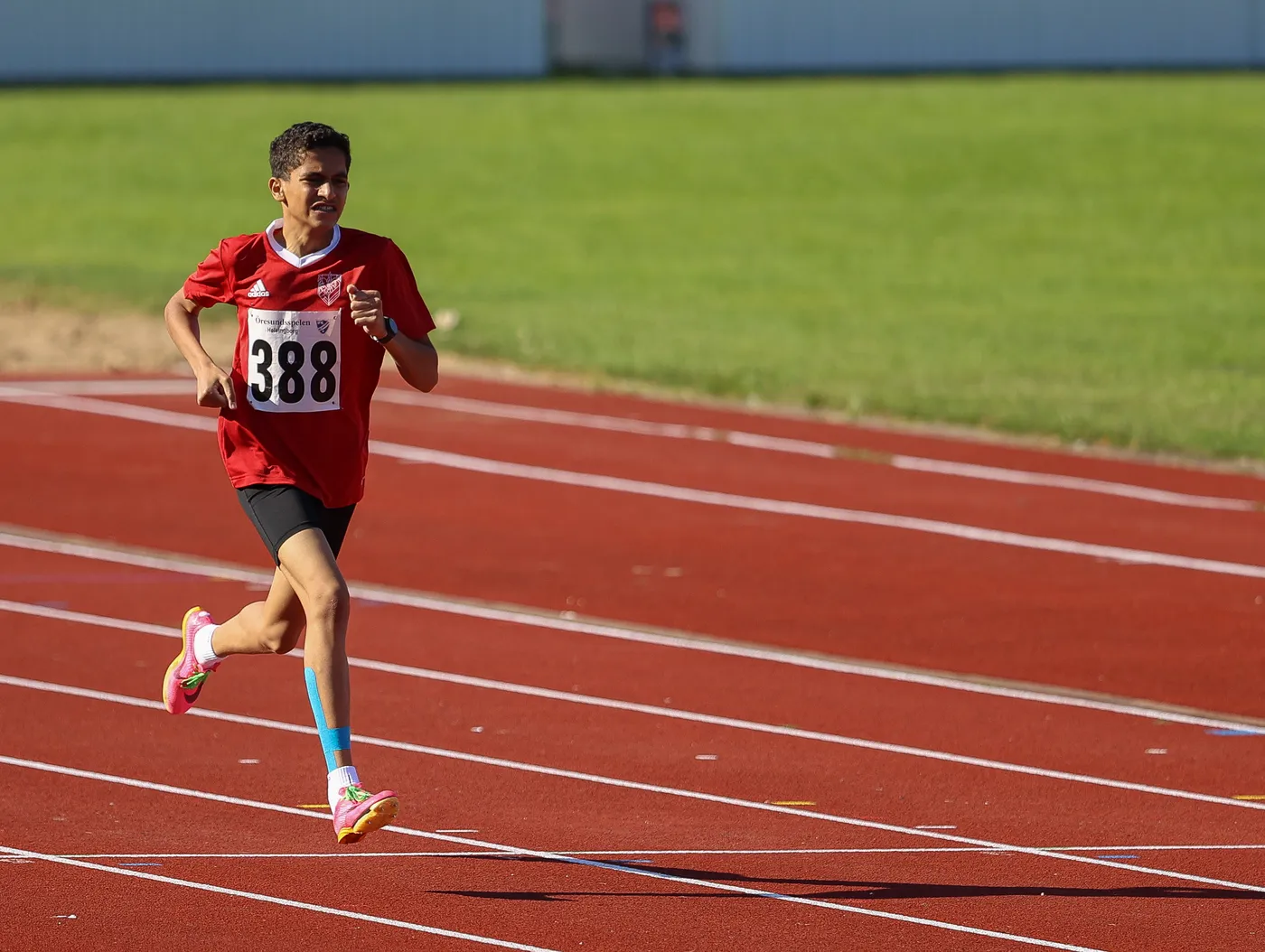 عداء يرتدي قميصًا أحمر يحمل الرقم 388 يسير بسرعة على مضمار رياضي ذي لون أحمر. الأرضية محاطة بالعشب الأخضر في الخلفية.