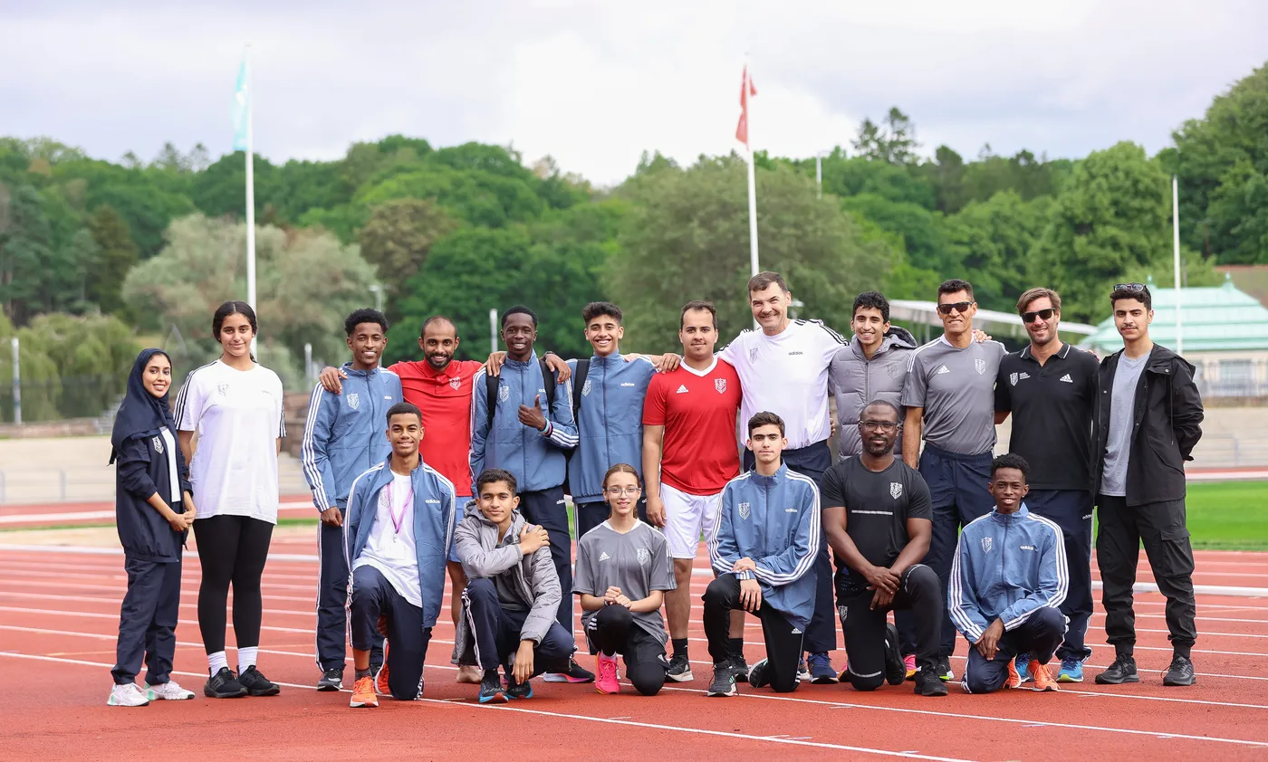 مجموعة من الشباب والبالغين يرتدون ملابس رياضية ويبتسمون معًا على مضمار رياضي. يتواجد بعضهم خلف الآخرين، بينما يقف آخرون أمامهم، مع الطبيعة الخضراء في الخلفية.