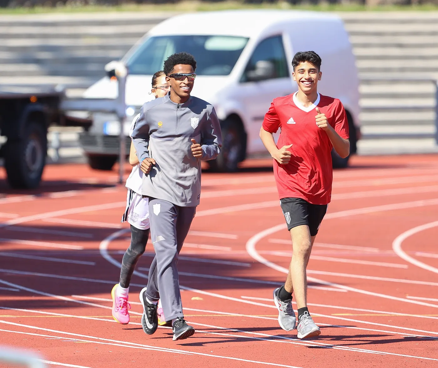 اثنان من المتسابقين يجريان على مضمار رياضي، حيث يرتدي أحدهم زي رياضي رمادي مع نظارات شمسية، بينما يرتدي الآخر قميصاً أحمر وشورت أسود. يظهر على وجههما الابتسامة وهما يستمتعان بالتدريب.