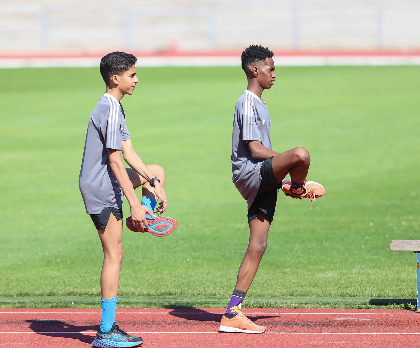شابان يتدربان على مضمار الجري، يقوم أحدهما برفع ساقه إلى الأعلى استعداداً للتمارين، بينما الآخر ينظر للأمام. يظهر كلاهما بملابس رياضية رمادية وقبعات.