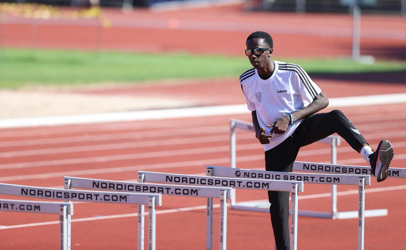 شخص يرتدي قميصاً رياضياً أبيض وسروالاً أسود، يتدرب على دفع ساقه فوق حواجز العدو في ميدان رياضي. يظهر في الخلفية مضمار رياضي مع حواجز مرتبة.