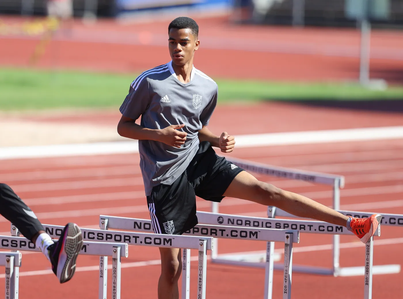 شاب يرتدي قميصًا رماديًا وشورتًا أسود، يتدرب في مضمار رياضي. يقوم برفع إحدى ساقيه فوق حاجز أثناء الجري.