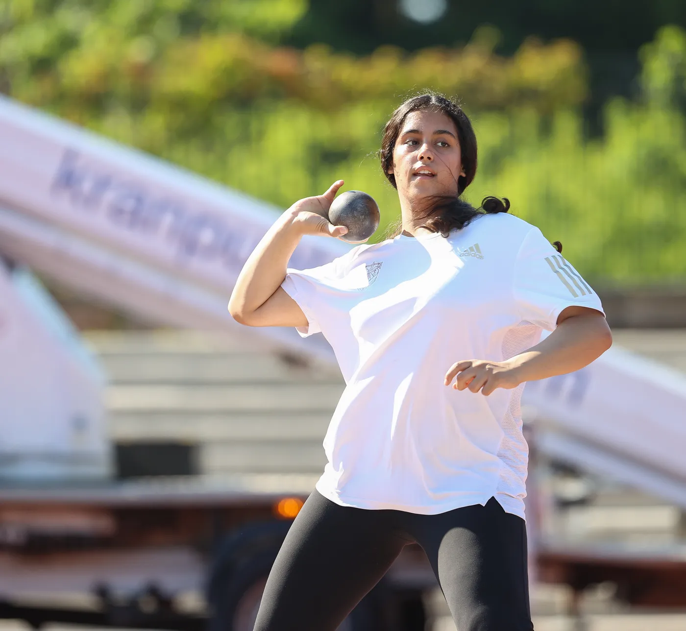 امرأة ترتدي قميصًا أبيض وسروالًا أسود، تستعد لرمي الكرة الثقيلة. تبدو مركزة وملتزمة أثناء التدريب، مع خلفية طبيعية تحتوي على مساحات خضراء ومعدات رياضية.