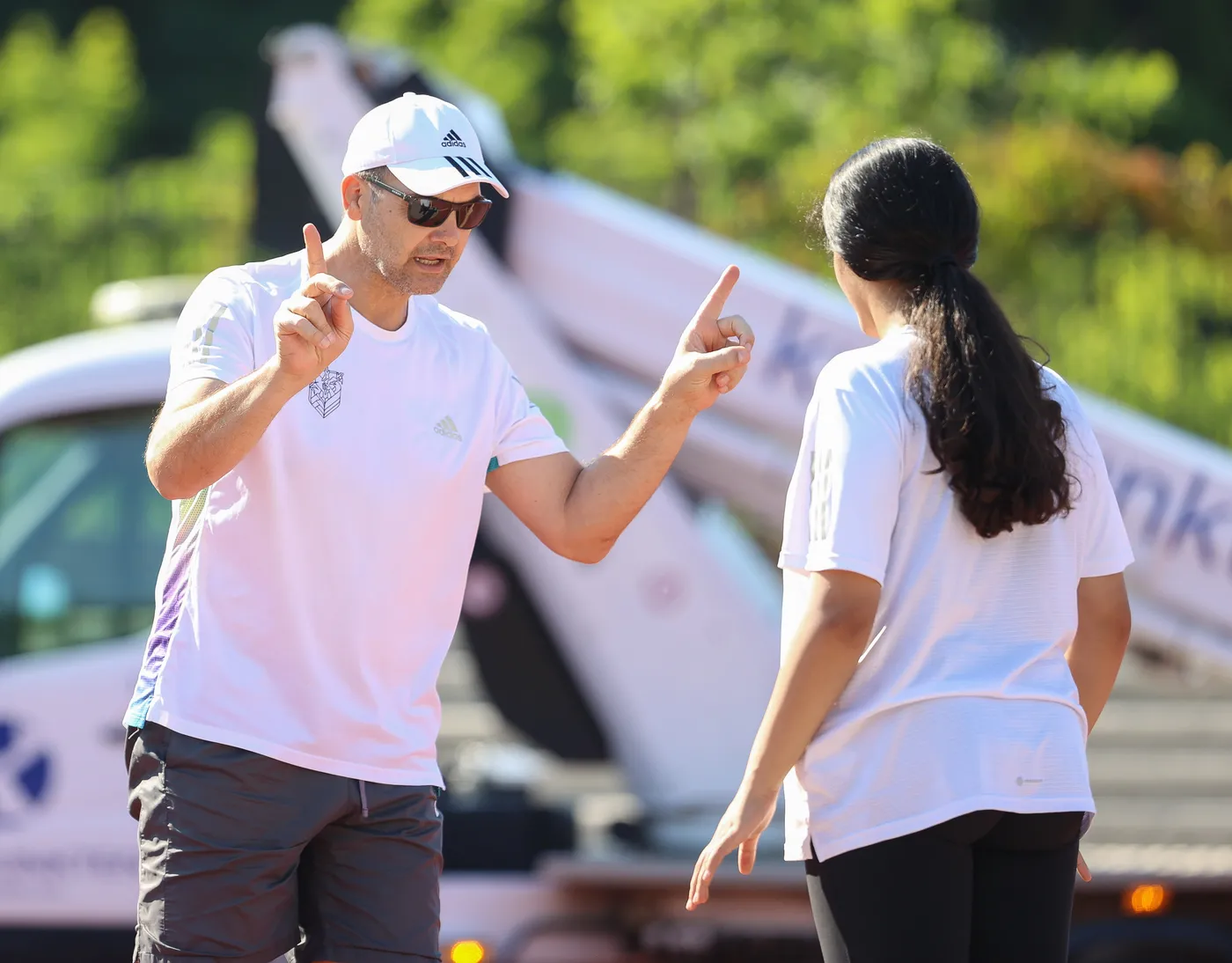 رجل يرتدي قبعة وساعة شمسية يقوم بإشارة بيده بينما يتحدث إلى امرأة ترتدي قميصًا أبيض. يبدو أن الحديث يدور حول التدريب أو النشاط الرياضي في الهواء الطلق، مع خلفية خضراء وأخرى لمركبة قربهما.