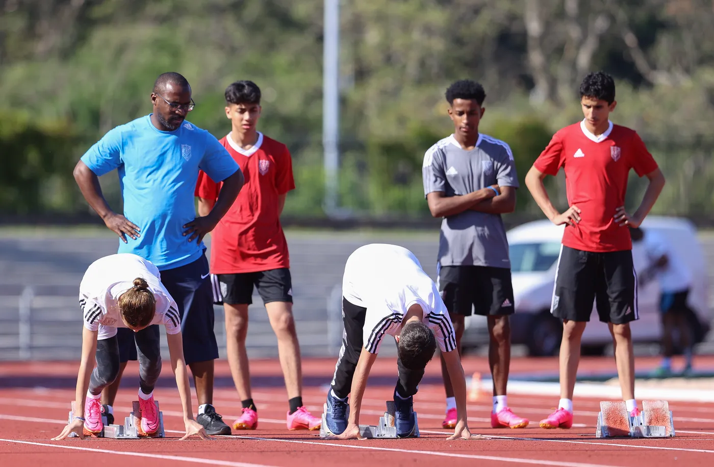 مدرب يشرف على مجموعة من العدائين الشباب أثناء التدريب على مضمار رياضي، حيث يتخذ بعض المتسابقين وضع الانطلاق بينما يتابع الآخرون عن كثب. يرتدون زيًا رياضيًا ويظهرون حماسًا للاستعداد للتدريب.