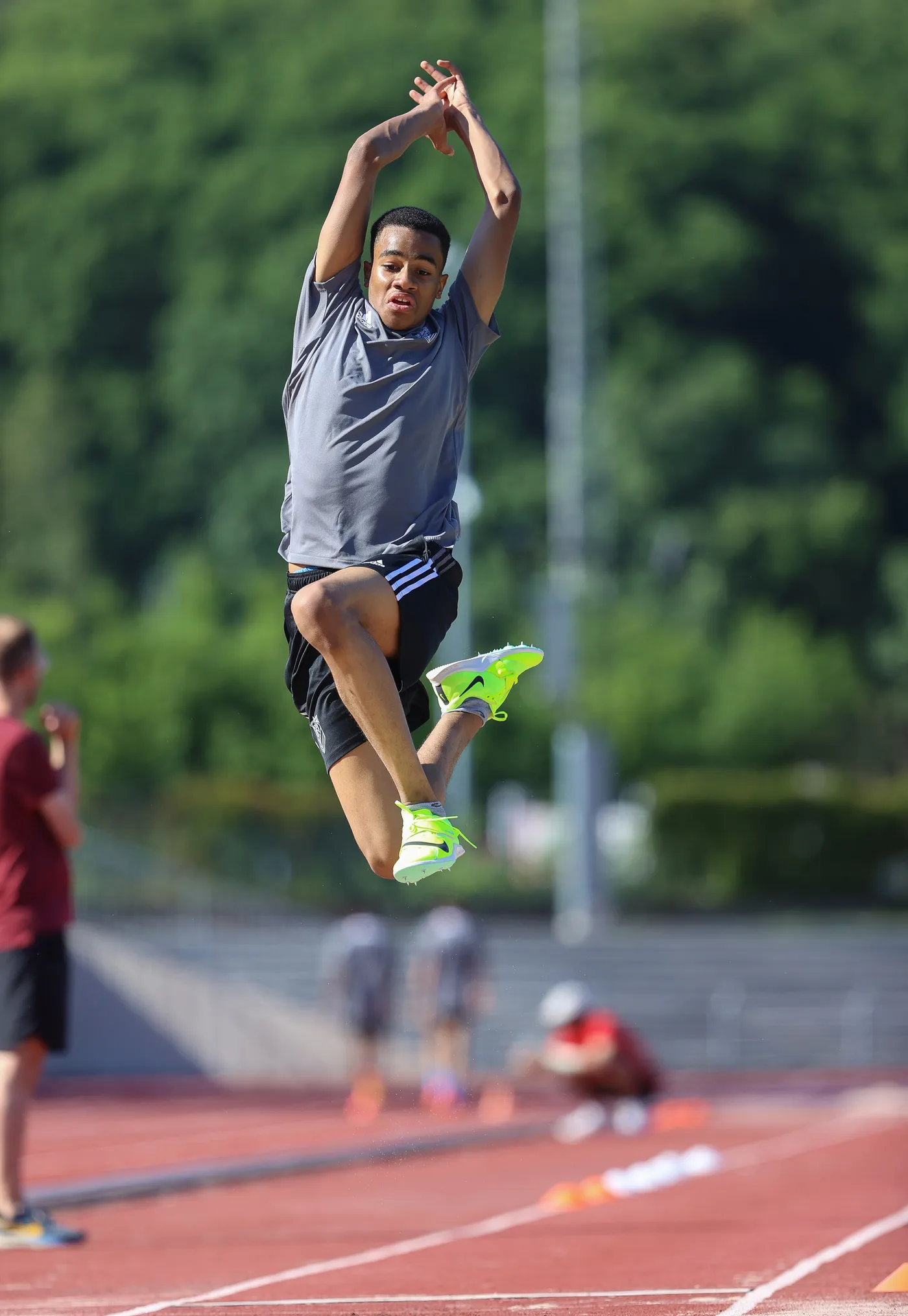 شاب يرتدي ملابس رياضية رمادية وسروال قصير أسود، يقوم بقفزة أثناء المنافسة في مضمار رياضي. تُظهر الصورة خلفية خضراء ومتناقضة مع المضمار الأحمر، مع عدد من الأشخاص الآخرين الذين يشاهدون الحدث من بعيد.