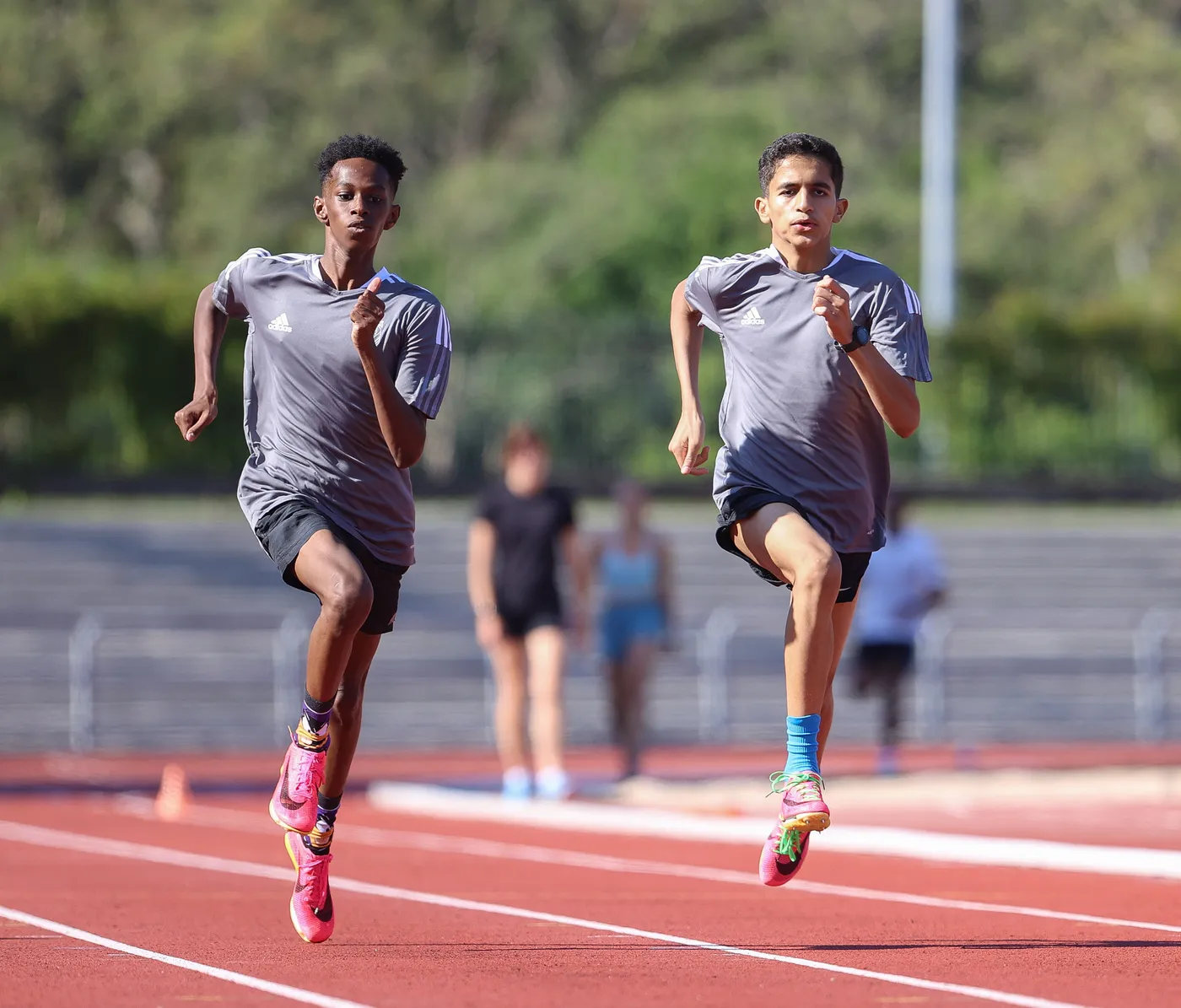 رجلان صغيران يركضان على مضمار رياضي بلون أحمر، يرتديان قمصاناً رمادية شورتات داكنة والأحذية ملونة. تظهر خلفهما مجموعة من العدائين الآخرين في الخلفية.