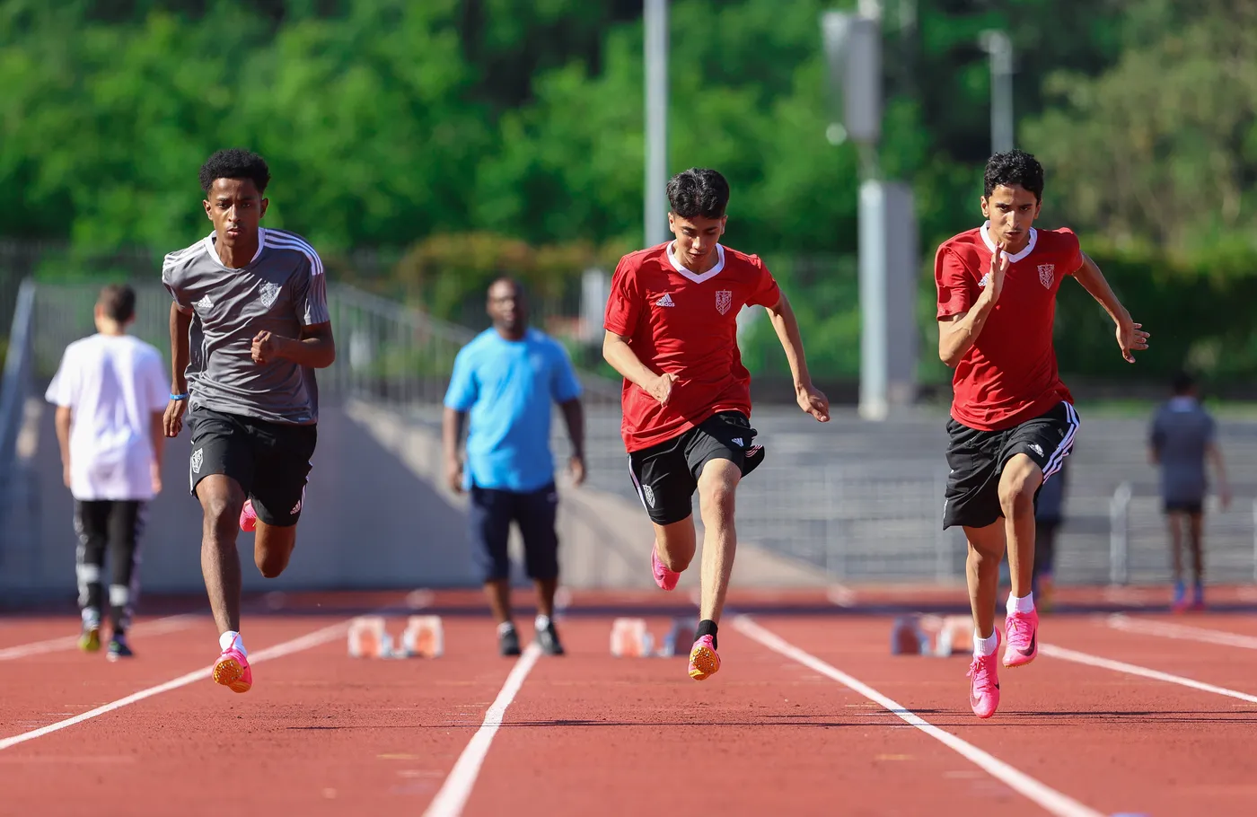 يظهر ثلاثة عدائين يرتدون قمصان حمراء يركضون على مضمار رياضي، مع وجود شخصين آخرين في الخلفية. يتواجد في الخلف أيضًا مدرب يرتدي قميصًا أزرق.