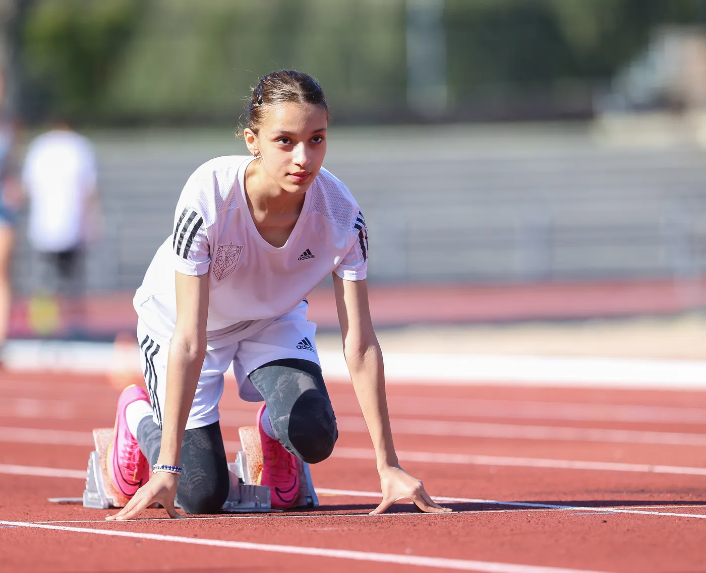 فتاة ترتدي قميصًا أبيض وسروالًا رياضيًا وهي تستعد للانطلاق في سباق عداء، تتخذ وضعية الانطلاق على المضمار الأحمر، بينما يبدو التركيز على وجهها. الخلفية تحتوي على متسابقين آخرين في مضمار تدريب.