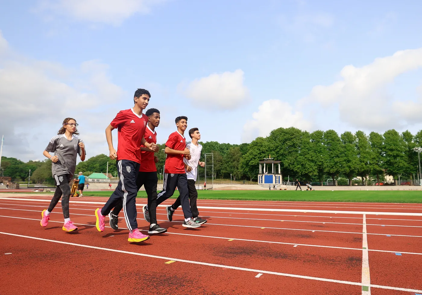 مجموعة من الشباب، تتكون من أربعة أولاد وبنت، يقومون بالجري على مضمار رياضي بلون أحمر. يرتدون قمصان حمراء وسراويل رياضية داكنة، وتظهر في الخلفية أشجار ومرافق رياضية.