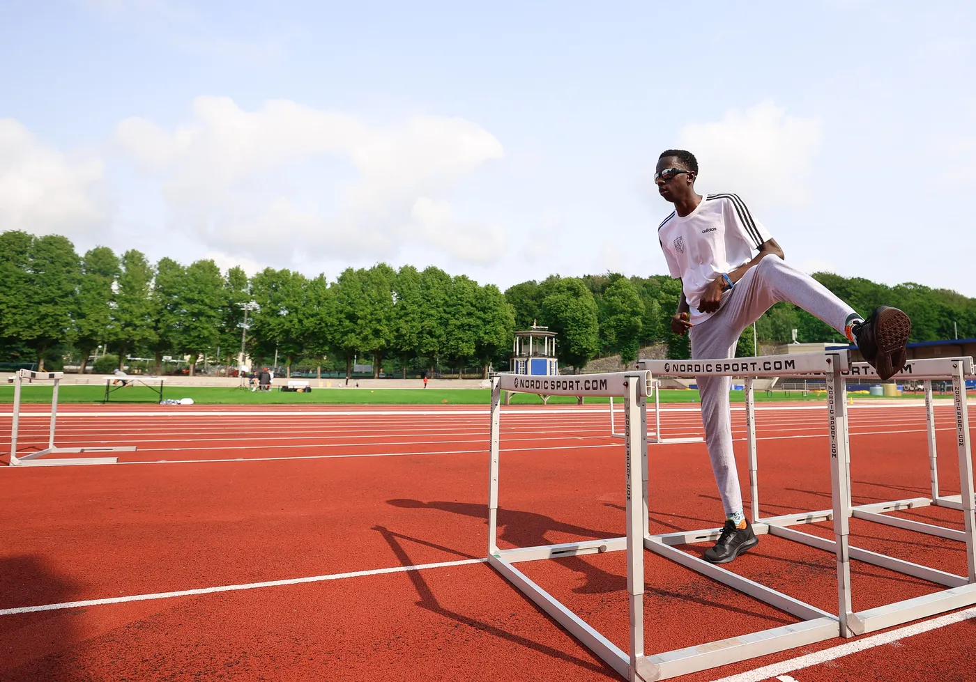 عداء يرتدي ملابس رياضية بيضاء وأسود يقف بجانب حواجز السباق، يستعد للانطلاق، بينما تظهر في الخلفية حلبة رياضية مع أشخاص آخرين يمارسون النشاط الرياضي. الأشجار الخضراء والطبيعة تحيط بالمكان.