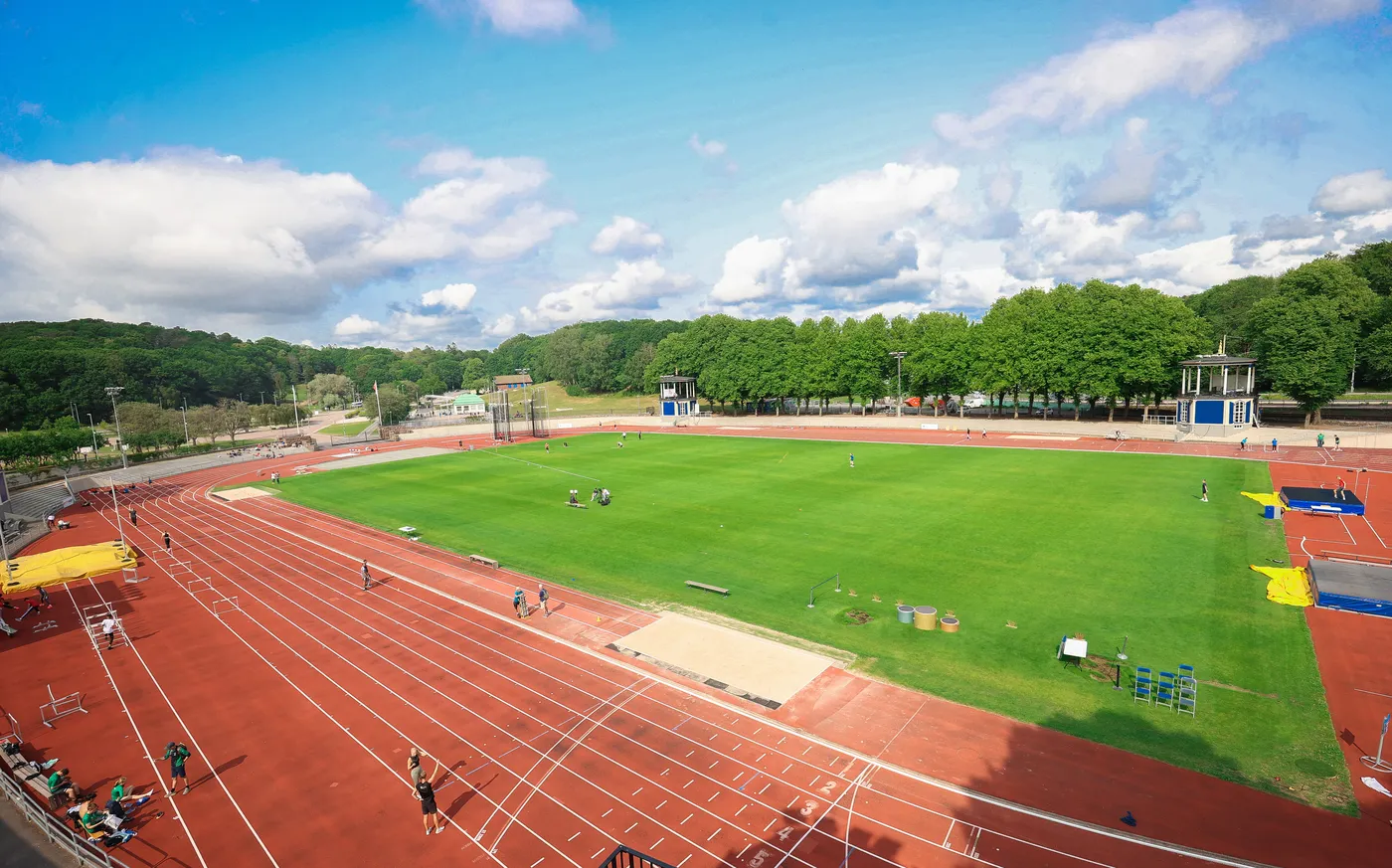 ملعب رياضي واسع يحتوي على مسارات تدريبية حمراء وحقل أخضر وسط الطبيعة، محاط بأشجار خضراء. تظهر مجموعة من الأشخاص يمارسون أنشطة رياضية مختلفة تحت سماء زرقاء مغطاة بسحب بيضاء.