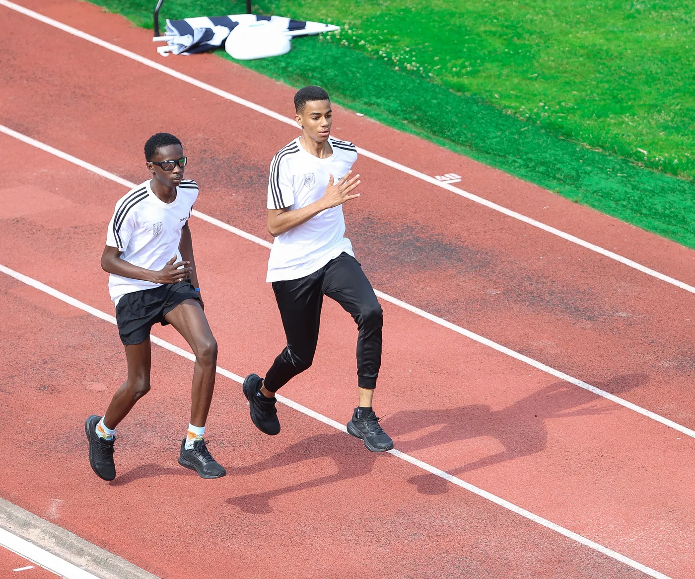 يوجد شابين يجريان على مضمار رياضي. يرتدي كلاهما قمصانا بيضاء مع خطوط سوداء وسراويل رياضية، حيث يبدو أن أحدهما يرتدي نظارات شمسية.