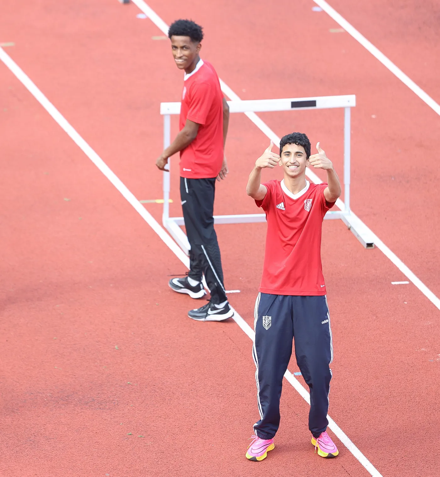 شاب يرتدي قميصًا أحمر وسروالًا داكنًا، يبتسم ويرفع إبهاميه في إشارة الإيجابية. خلفه، شاب آخر أيضاً يرتدي قميصًا أحمر، يبدو أنه يستعد لأحد التمارين الرياضية على مضمار ملون باللون الأحمر.