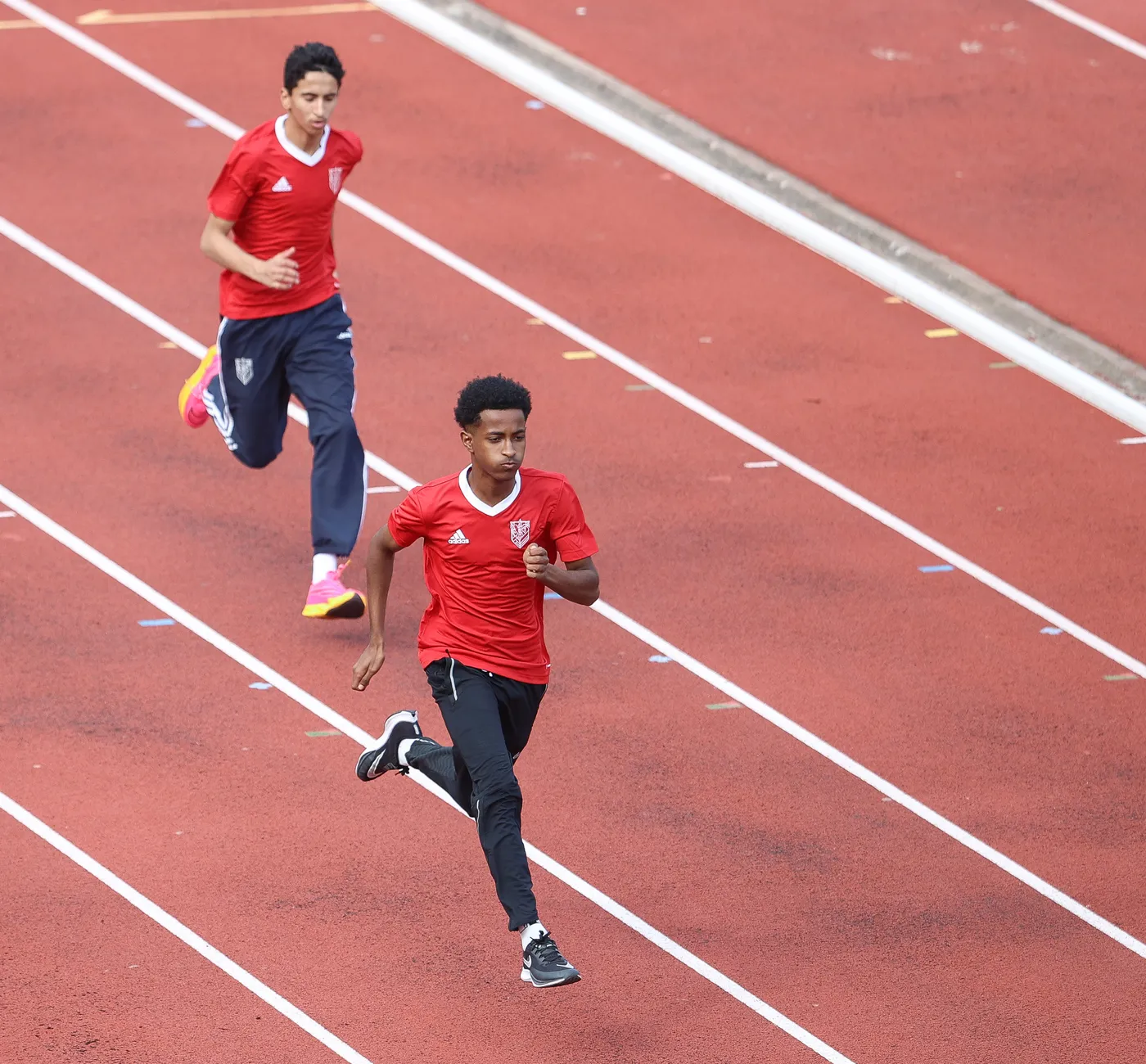 عداء يرتدي قميصًا أحمر يتقدم على مضمار سباق، بينما عداء آخر يقترب منه من الخلف. الأرضية حمراء وموجهة لسباق الجري.