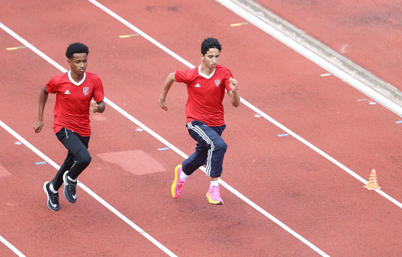 يشارك شابان في سباق على مضمار رياضي، يرتديان قمصانًا حمراء وسراويل رياضية. أحدهما يرتدي حذاءً ورديًا، بينما الآخر في حذاء أسود، ويبدوان مركزين في السباق.