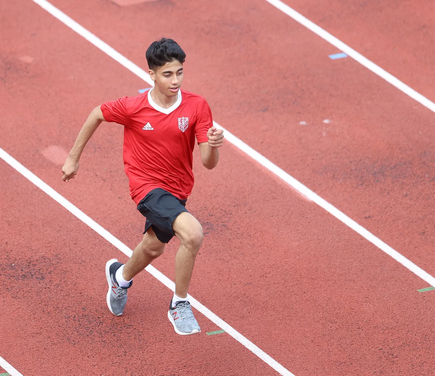 شاب يرتدي قميصًا أحمر ويركض على مضمار رياضي ذو لون أحمر، يظهر في منتصف حركة الركض. يمسك بيده اليمنى بها عضلة مشدودة، وتبدو على وجهه تعبيرات التركيز والحماس.