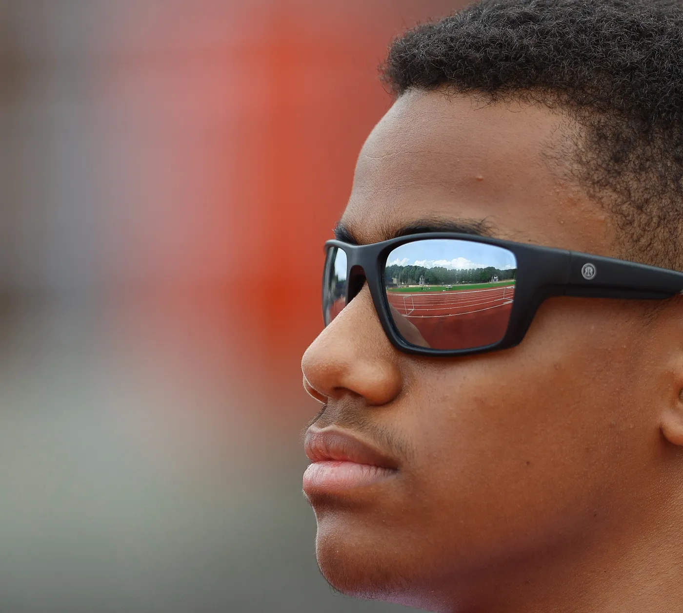 شاب يرتدي نظارات شمسية، يظهر بوضوح في عينيه انعكاس لمضمار رياضي وأشخاص يتدربون. مع خلفية مليئة بالألوان الدافئة، يتميز بالتعبير الجاد والتركيز.