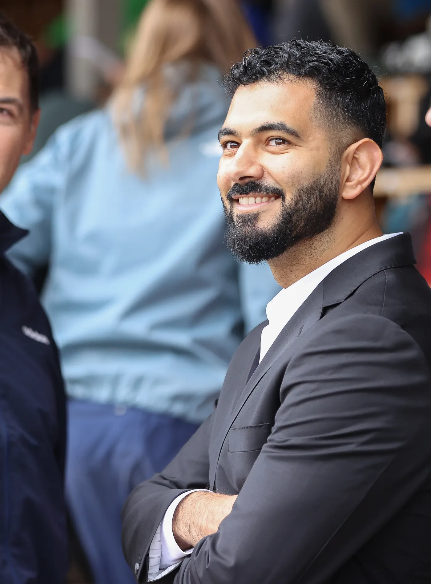 رجل ذو لحية وشعر مقصوص بشكل أنيق، يرتدي بدلة سوداء وقميص أبيض، مبتسم بينما يقف مع ذراعيه متقاطعتين. يوجد أشخاص آخرون في الخلفية، مما يدل على حدث اجتماعي.