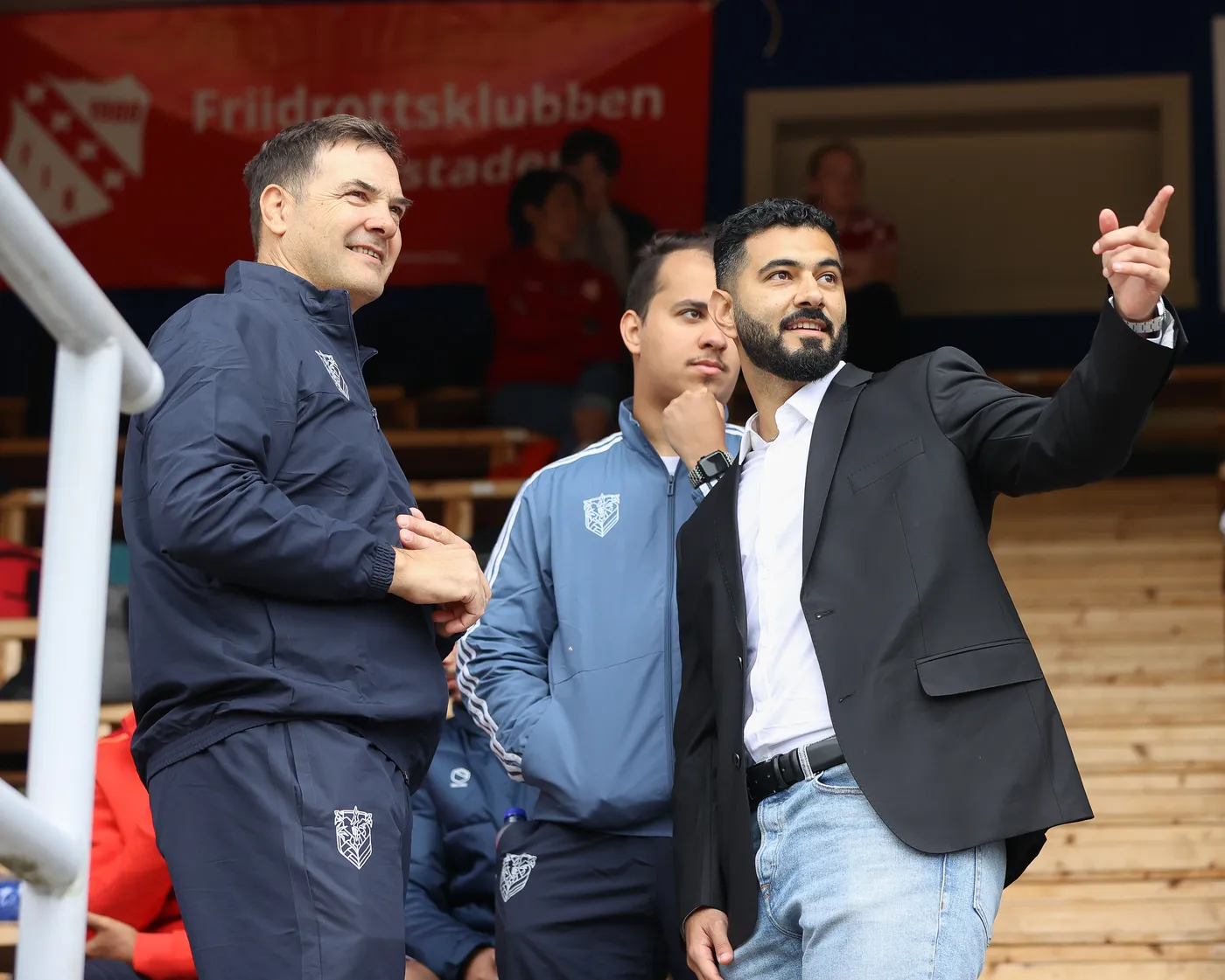 رجلان يقفان في مدرج رياضي، أحدهما يرتدي زي رياضي والآخر بدلة سوداء، يتبادلان الحديث بينما يشير الرجل في البدلة بإصبعه نحو جهة معينة. يوجد شاب ثالث خلفهما يبدو متفحصًا وضعه.