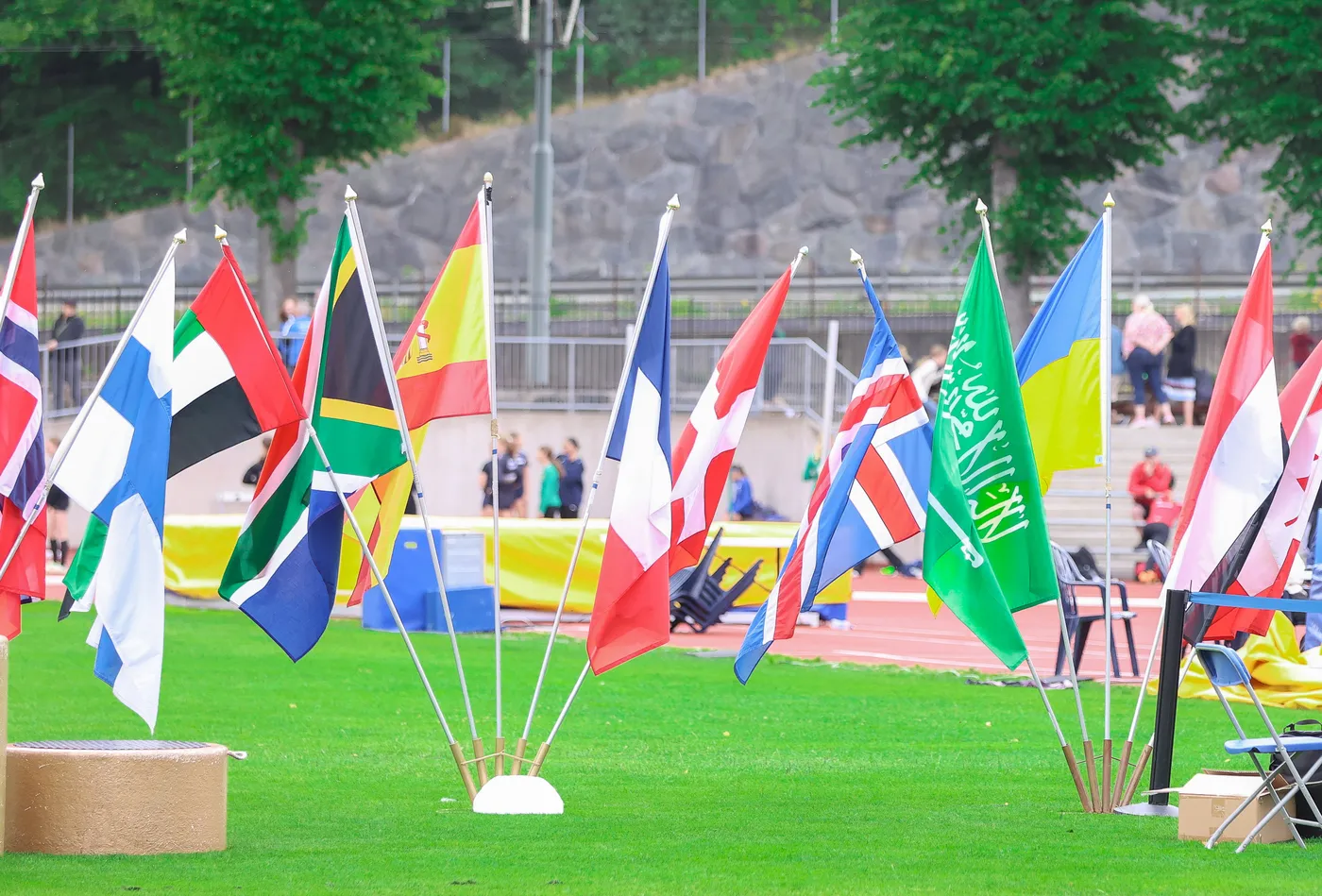 تتواجد مجموعة من الأعلام تُرفرف في مكان مفتوح، تمثل عدة دول مختلفة، بما في ذلك الأعلام البريطانية، والألمانية، والجنوب أفريقية، والسعودية. يحيط بالأعلام ملعب رياضي وأشخاص في الخلفية.