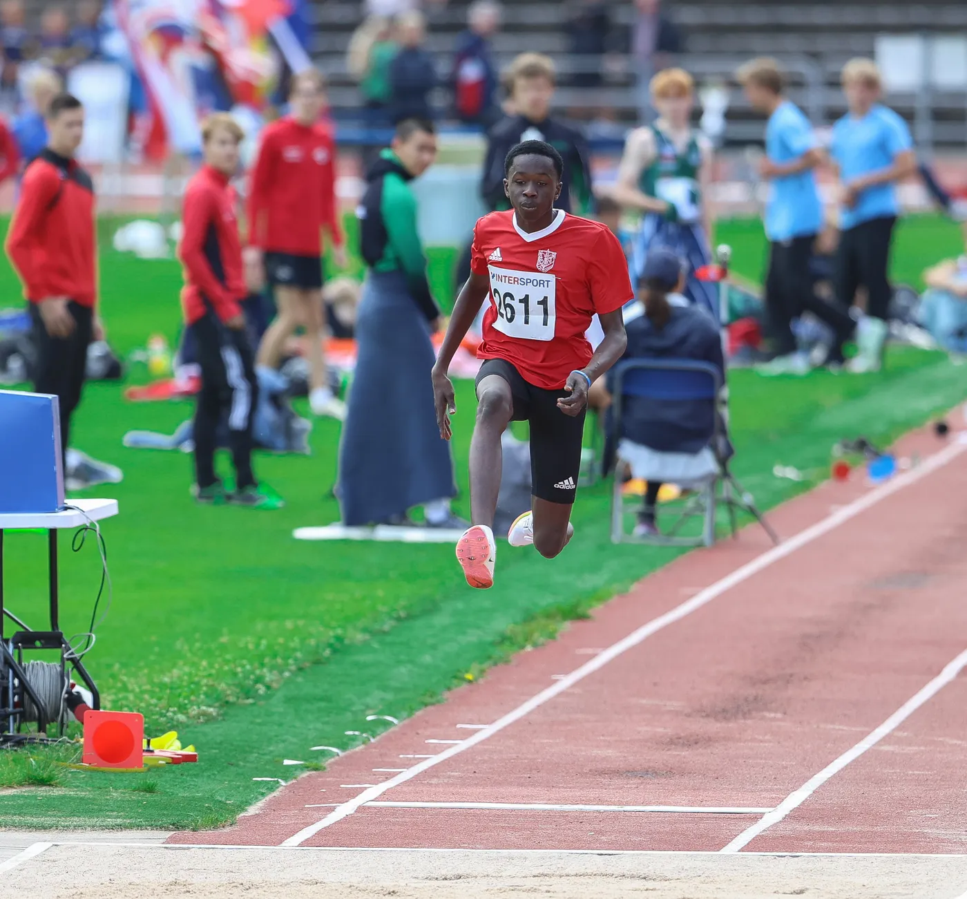 يظهر عداء في الهواء وهو يؤدي قفزة طويلة خلال مسابقة في ملعب رياضي، مرتديًا قميصًا أحمر مع رقم ١١. في الخلفية، يوجد عدد من المتسابقين والمشجعين يجلسون على المقاعد.