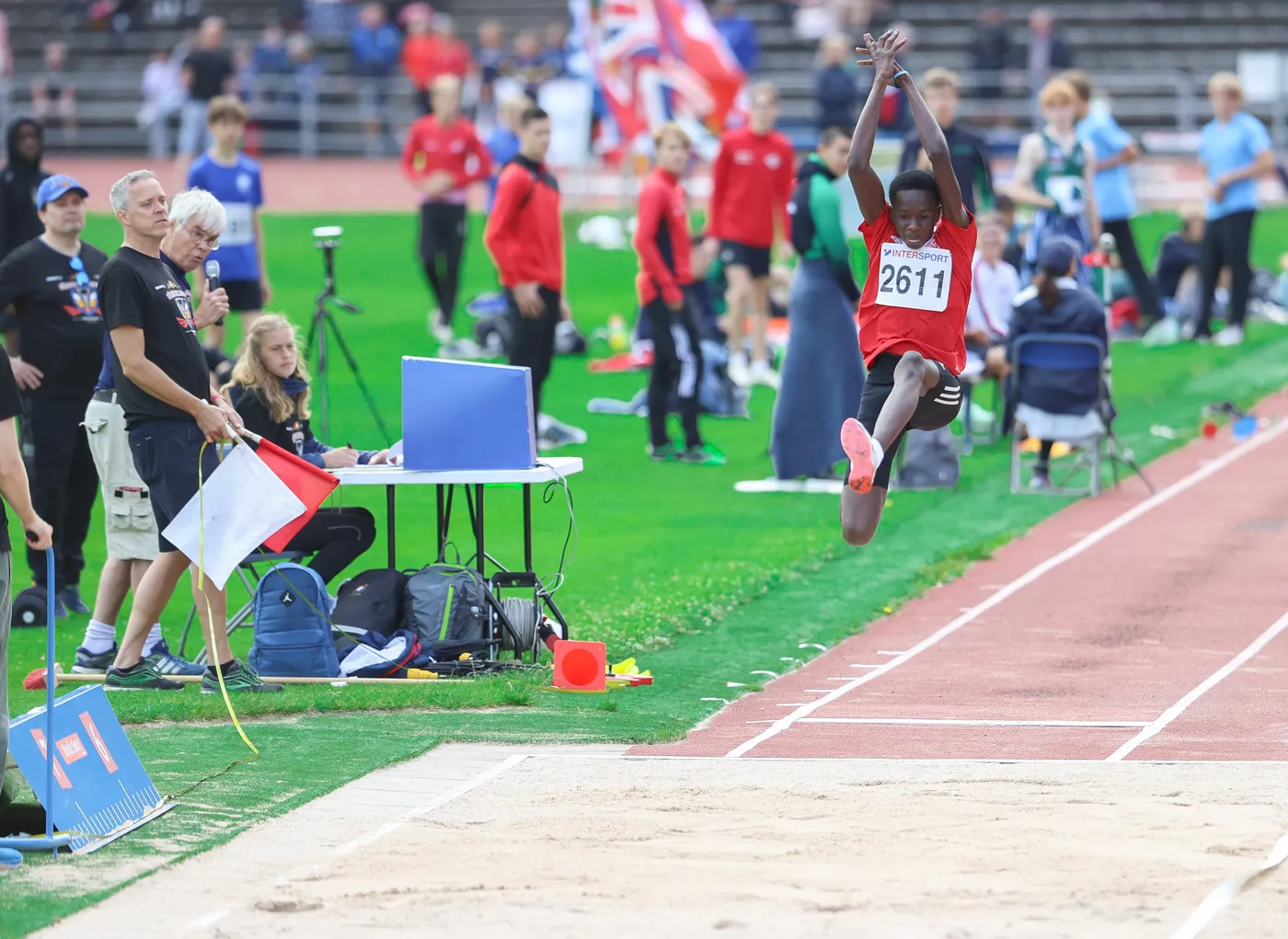 عداء يقفز في مسابقة القفز الطويل خلال حدث رياضي، يرتدي تيشيرتًا أحمر ورقمًا على صدره. في الخلفية، يوجد حكام ومشجعون، مع أعلام ملونة ترفرف.
