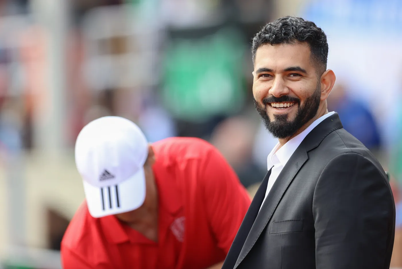 رجل مبتسم ذو لحية وملابس رسمية سوداء يقف في المقدمة، بينما يتم تقديم المساعدة من قبل شخص آخر يرتدي قميصًا أحمر وقبعة بيضاء. الخلفية تظهر حشودًا ومشاهد رياضية.