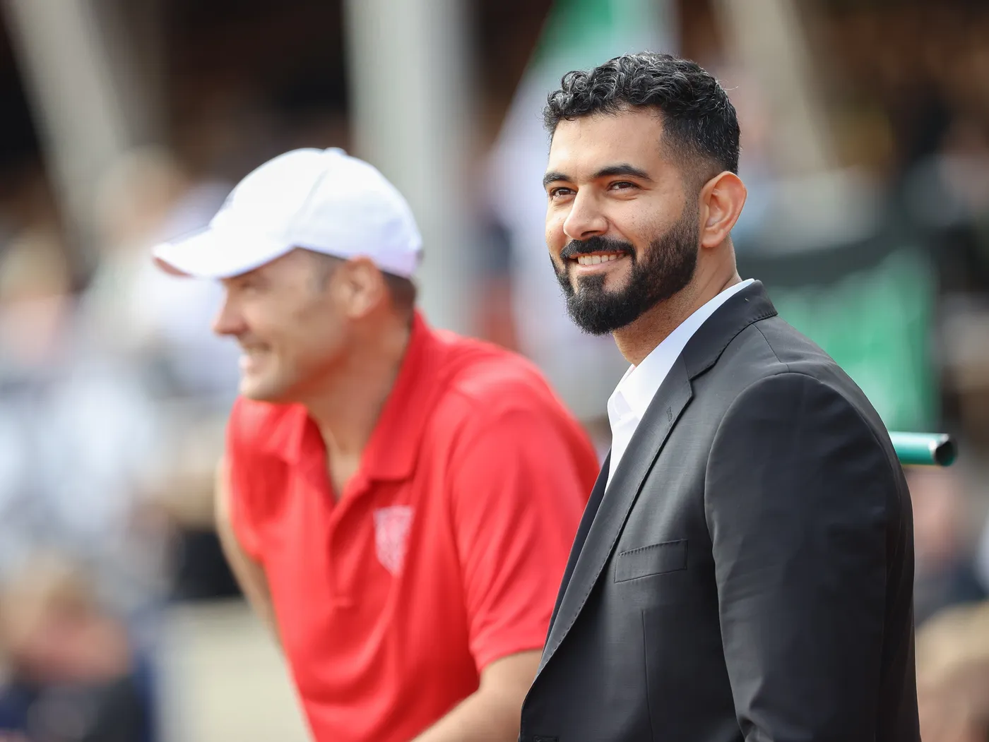 رجل يرتدي بدلة سوداء وقميصًا أبيض يبتسم ويبدو سعيدًا، بينما يقف بجانبه رجل آخر يرتدي قميصًا أحمر وقبعة بيضاء. تظهر في الخلفية مجموعة من الأشخاص ومكان ينظم فيه حدث.