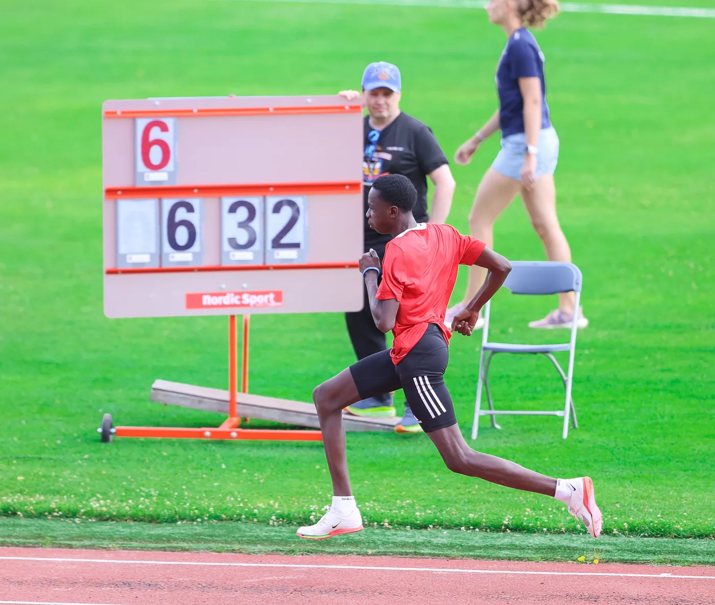 عداء يرتدي قميصًا أحمر وشورتًا أسود يجري على المضمار، خلفه لوحة تسجيل تعرض الأرقام 6 و32. في الخلفية، يوجد شخصان يتابعان الأحداث، مع خلفية من العشب الأخضر.