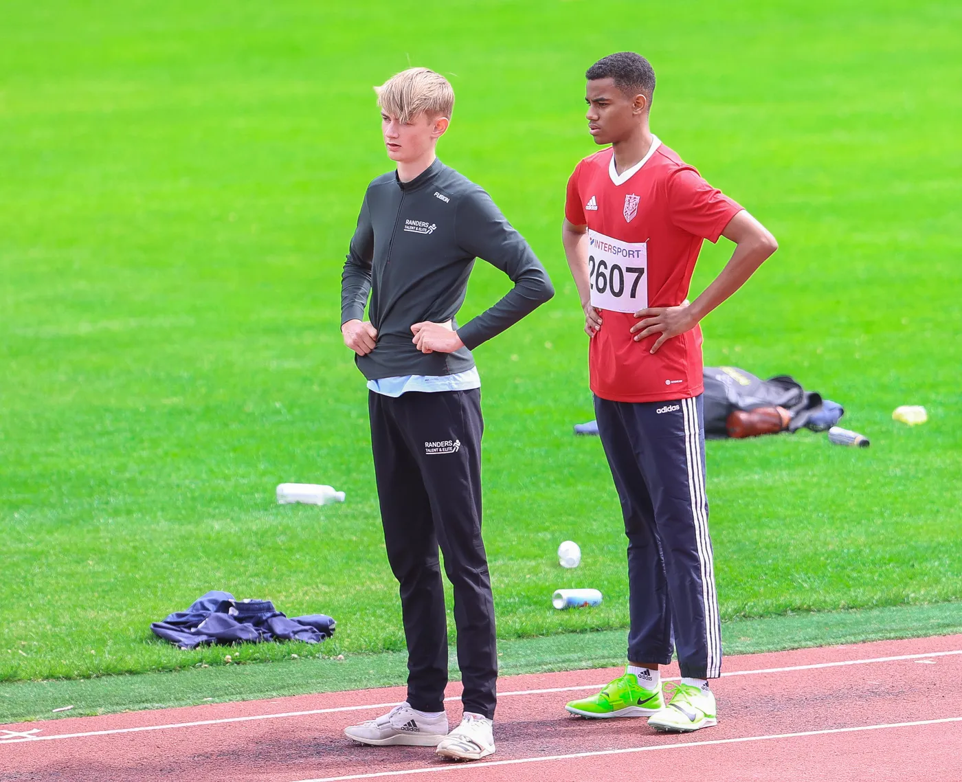 شابان يقفان على مضمار رياضي، يرتدي أحدهم قميصًا أخضر وسروالًا أسود، بينما يرتدي الآخر قميصًا أحمر ورقمًا على صدره. يظهر أمامهما عشب أخضر ومقاعد رياضية وأشياء متنوعة ملقاة على الأرض.