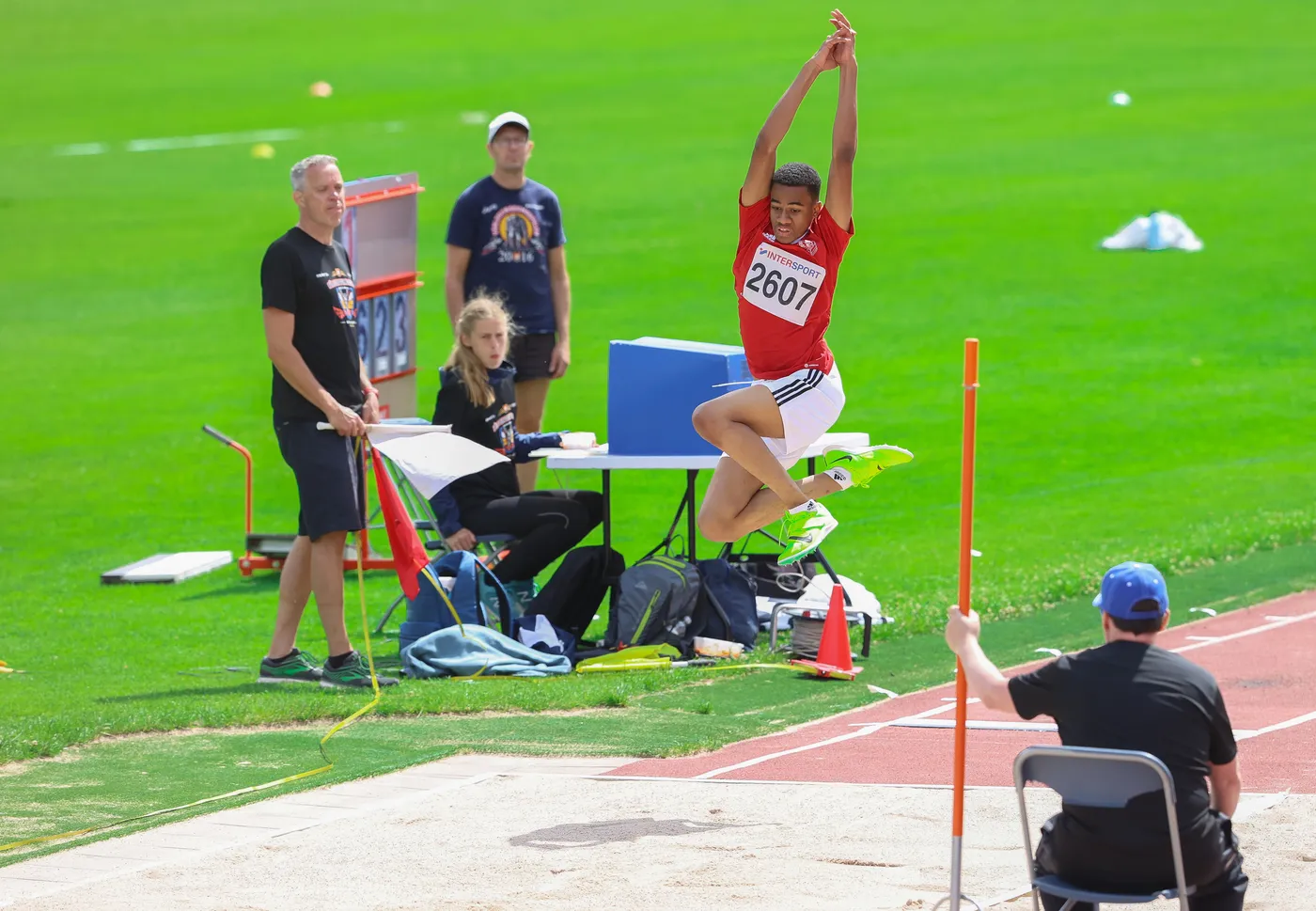 عداء في زي رياضي أحمر يقفز عالياً خلال منافسة رياضية، بينما يراقبه مجموعة من المدربين والمشجعين. في الخلفية، توجد معدات رياضية وأعلام ملونة، مع حقل أخضر واسع في المشهد.