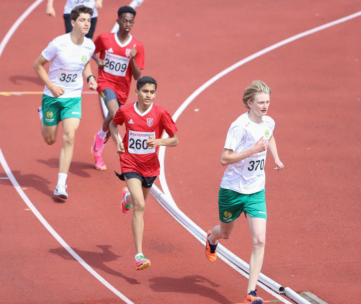 مجموعة من أربعة متسابقين على مضمار سميك باللون الأحمر. يرتدي أحد المتسابقين قميصًا أحمر ورقم 260، بينما يرتدي الآخرون قمصانًا بيضاء وخضراء. يتضح من ملامحهم ونشاطهم أنهم يتنافسون في سباق.