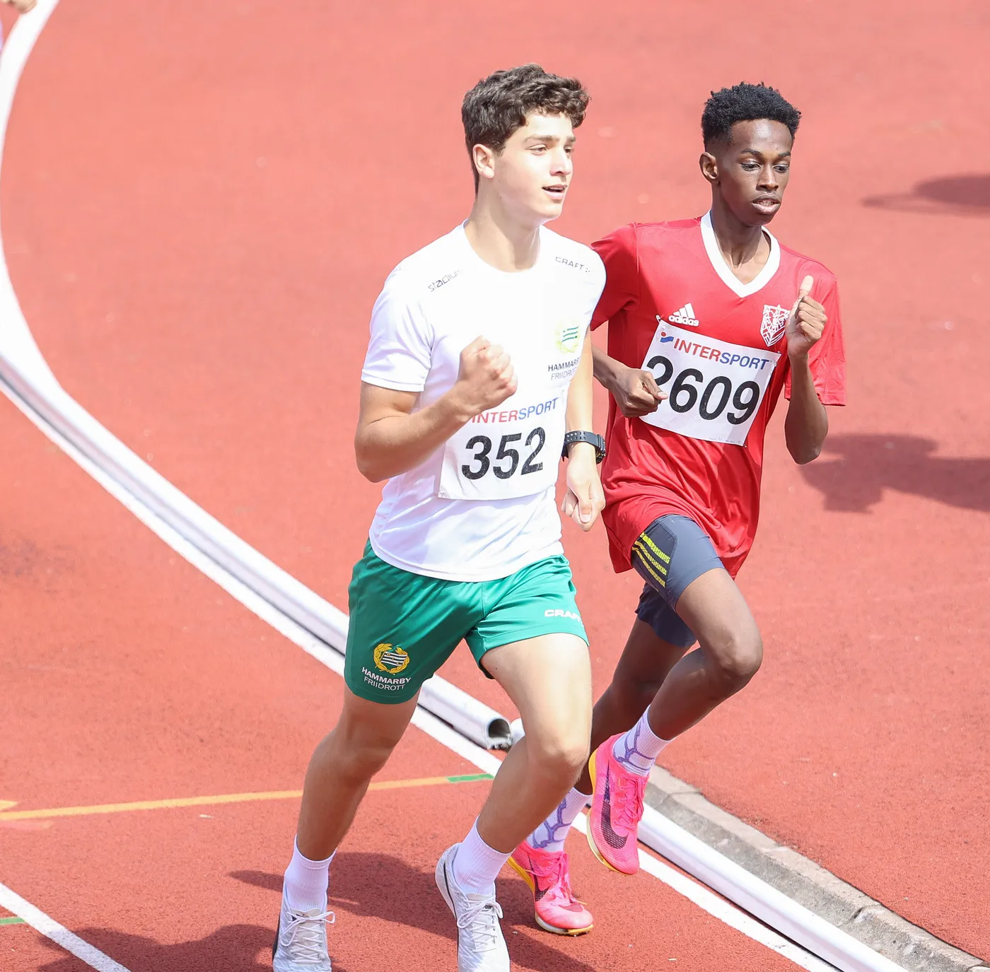 يشارك عدّاءان في سباق على مضمار رياضي. الأول يرتدي قميصًا أبيض وسروالًا أخضر، بينما الثاني يرتدي قميصًا أحمر وسروالًا داكنًا.