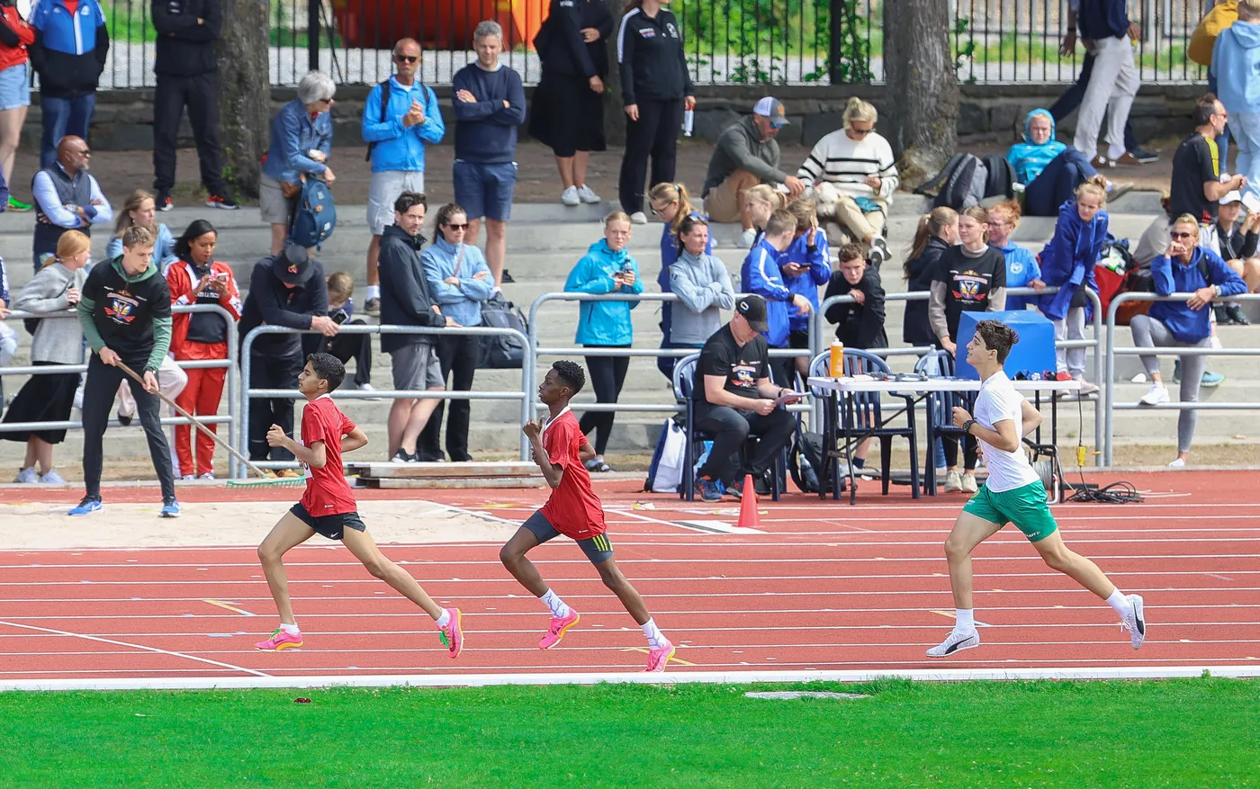 يظهر ثلاثة عدائين في سباق على مضمار رياضي، حيث يرتدي العداؤون قمصانًا حمراء وبيضاء. الجمهور يتابع من الخلف، يتضمن المشهد مجموعة متنوعة من الأشخاص. في الأمام، العداؤون يتنافسون بحماسة.