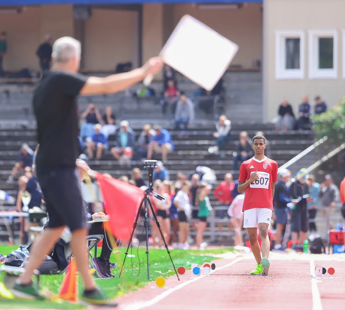 عداء في زي رياضي أحمر يحمل رقم 2607 يتقدم على مضمار التدريب، بينما يبدو حكم أو مسؤول في الخلف وهو يلوح بشيء. تجمع من المتفرجين يظهر في الخلفية مع وجود معدات تصوير ومؤشرات ملونة على الأرض.
