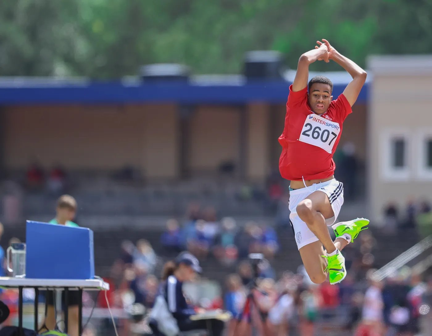 شاب يرتدي قميصًا أحمر وشورت أبيض يقوم بقفزة في مسابقة رياضية، مع رقم 2607 على صدره. في الخلفية، تظهر حشود من المتفرجين ومرافق رياضية.